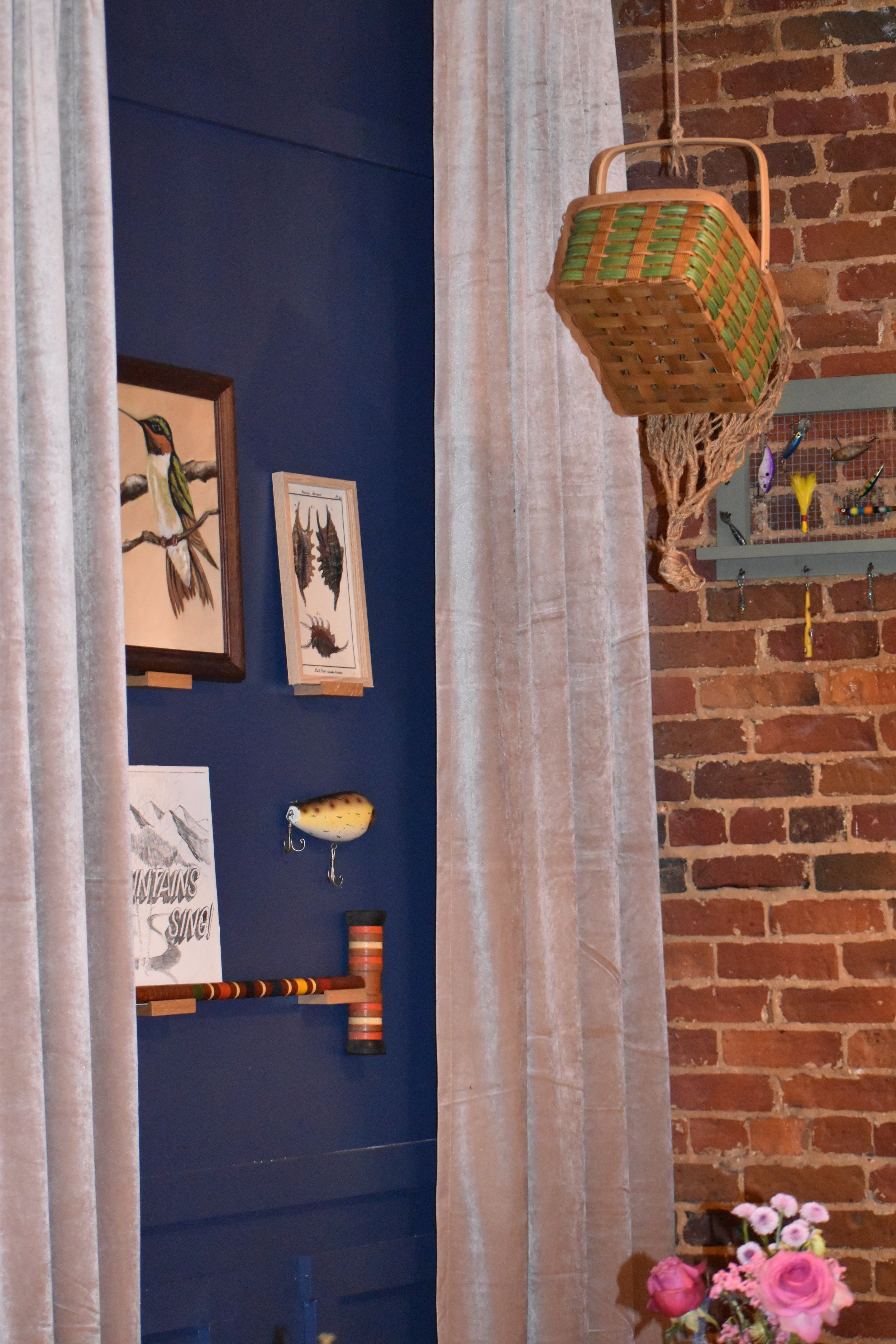 Interior of a room with a brick wall, beige curtains, and various bird-themed decorations, including framed bird illustrations, a small bird sculpture, and a basket hanging from the ceiling with a decorative net. There are also flowers in the bottom 