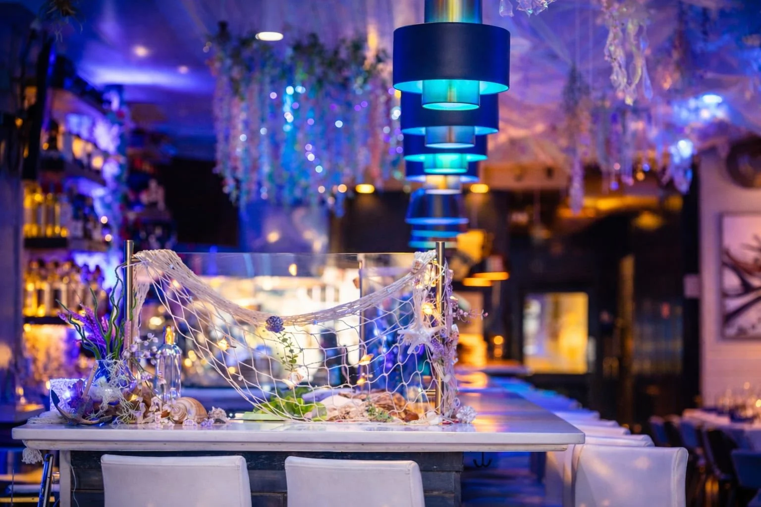 A decorated table with seashells, flowers, and a netted decoration inside a restaurant with blue hanging lamps and purple hanging streamers. Aquatic themed restaurant decor.