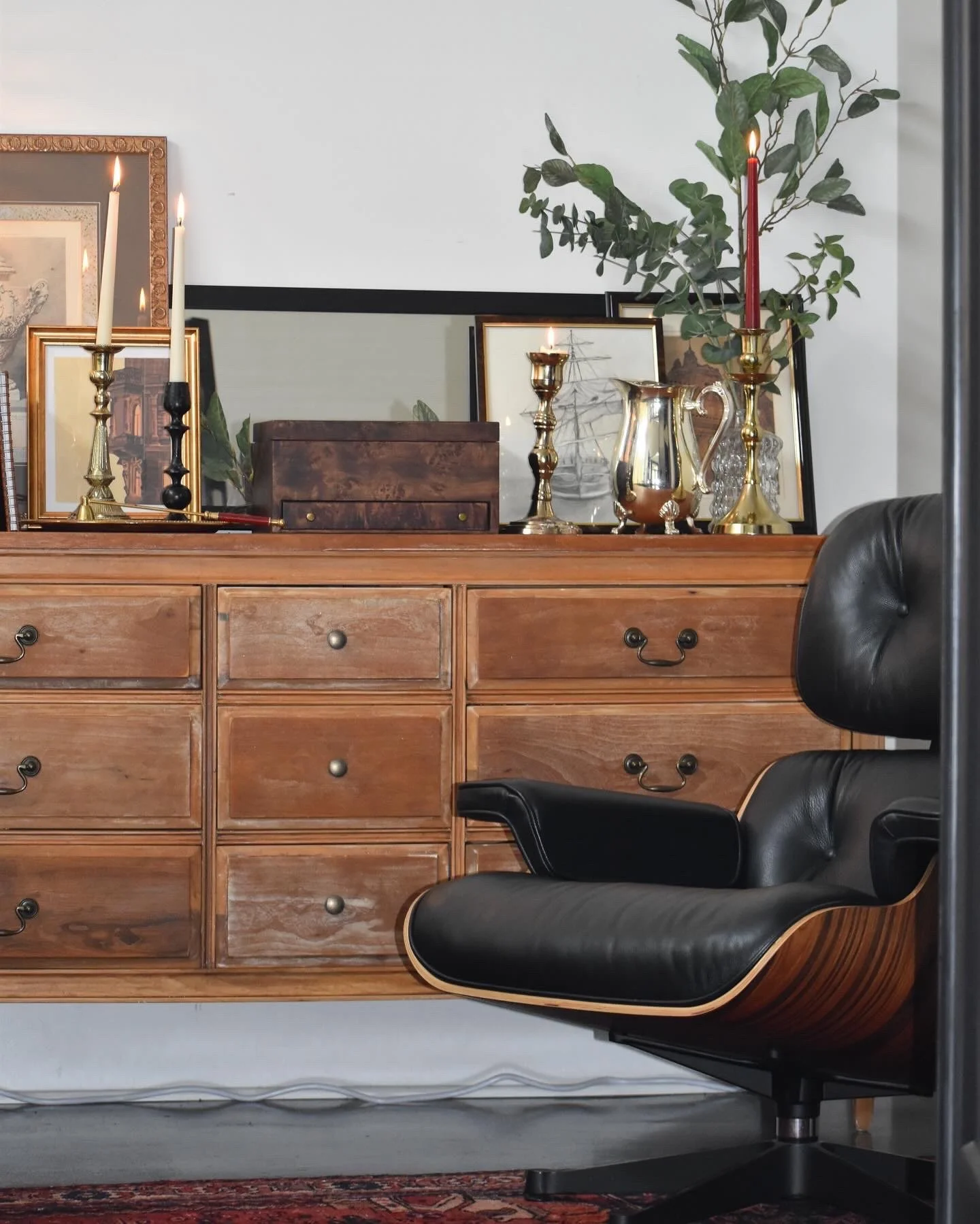 A wooden dresser decorated with framed artwork, candlesticks, a silver pitcher, and a potted plant in the background, with a black leather and wood lounge chair in the foreground.