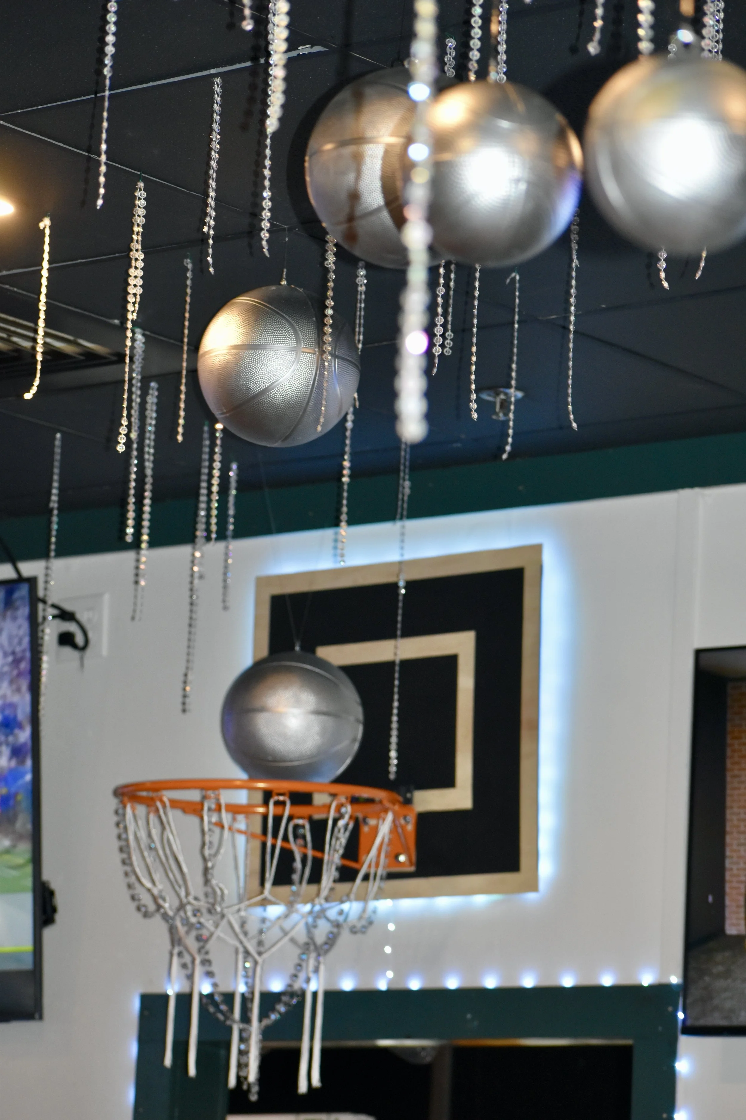 Indoor basketball-themed decor with silver basketballs hanging from the ceiling, a mini basketball hoop, and string lights. March Madness