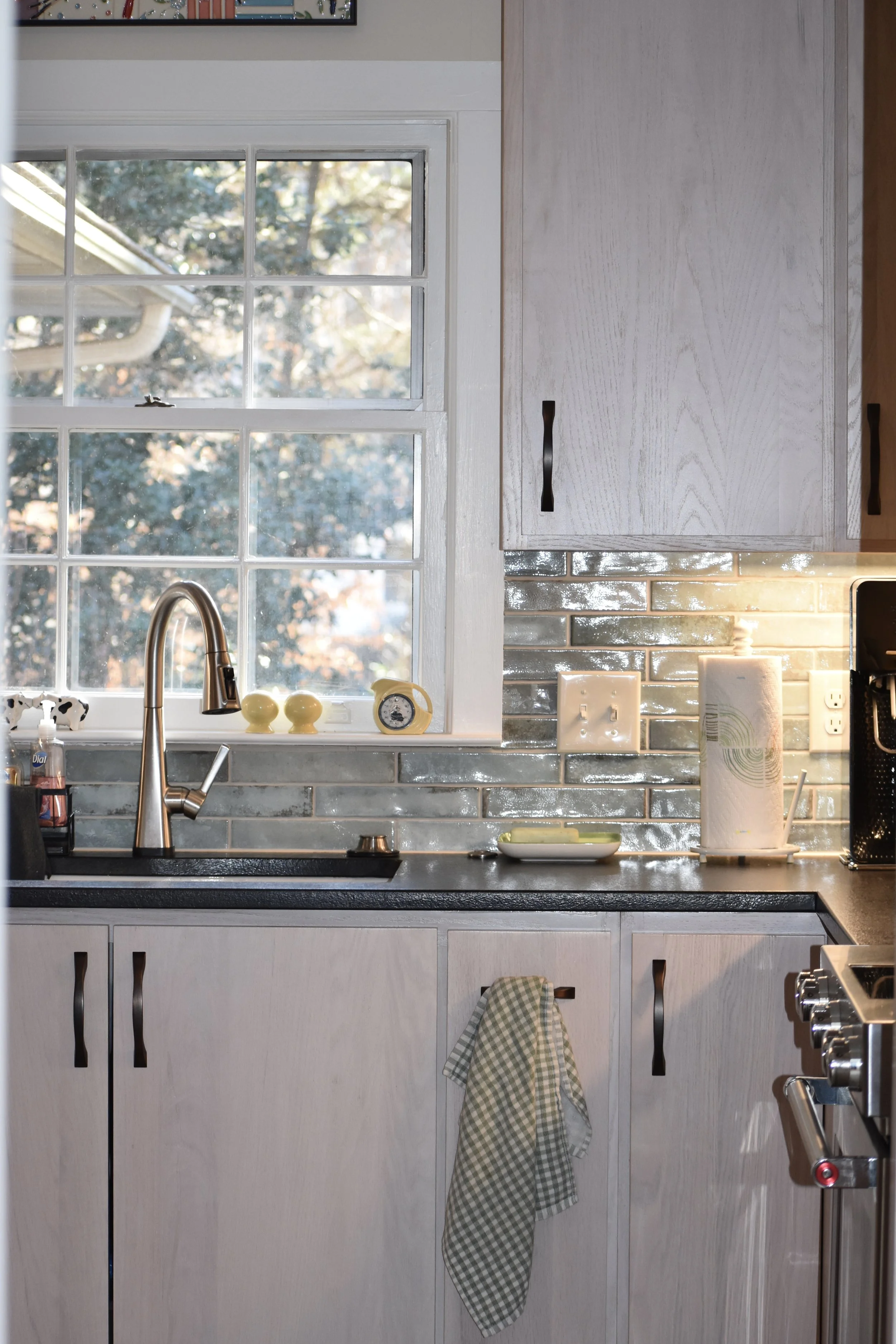 Kitchen with a window above the sink, a silver faucet, soap dispenser, kitchen paper roll, and various kitchen items on the black countertop; light-colored cabinets with black handles, a checkered cloth hanging from one handle, and a tiled backsplash