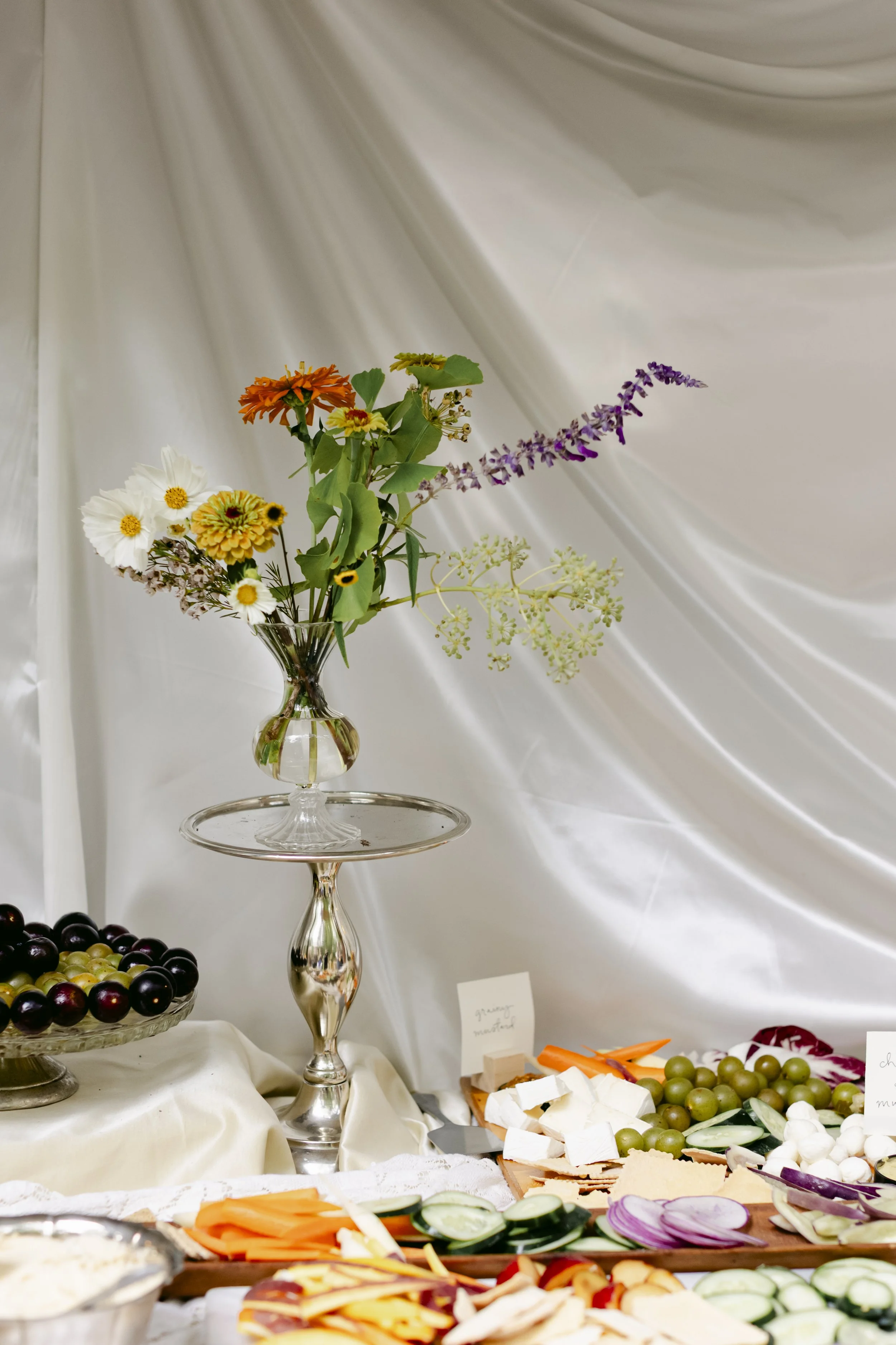 A vase with colorful flowers on a silver stand, placed on a table with fruits and vegetables, with a white draped background. Wedding florals around charcuterie board.