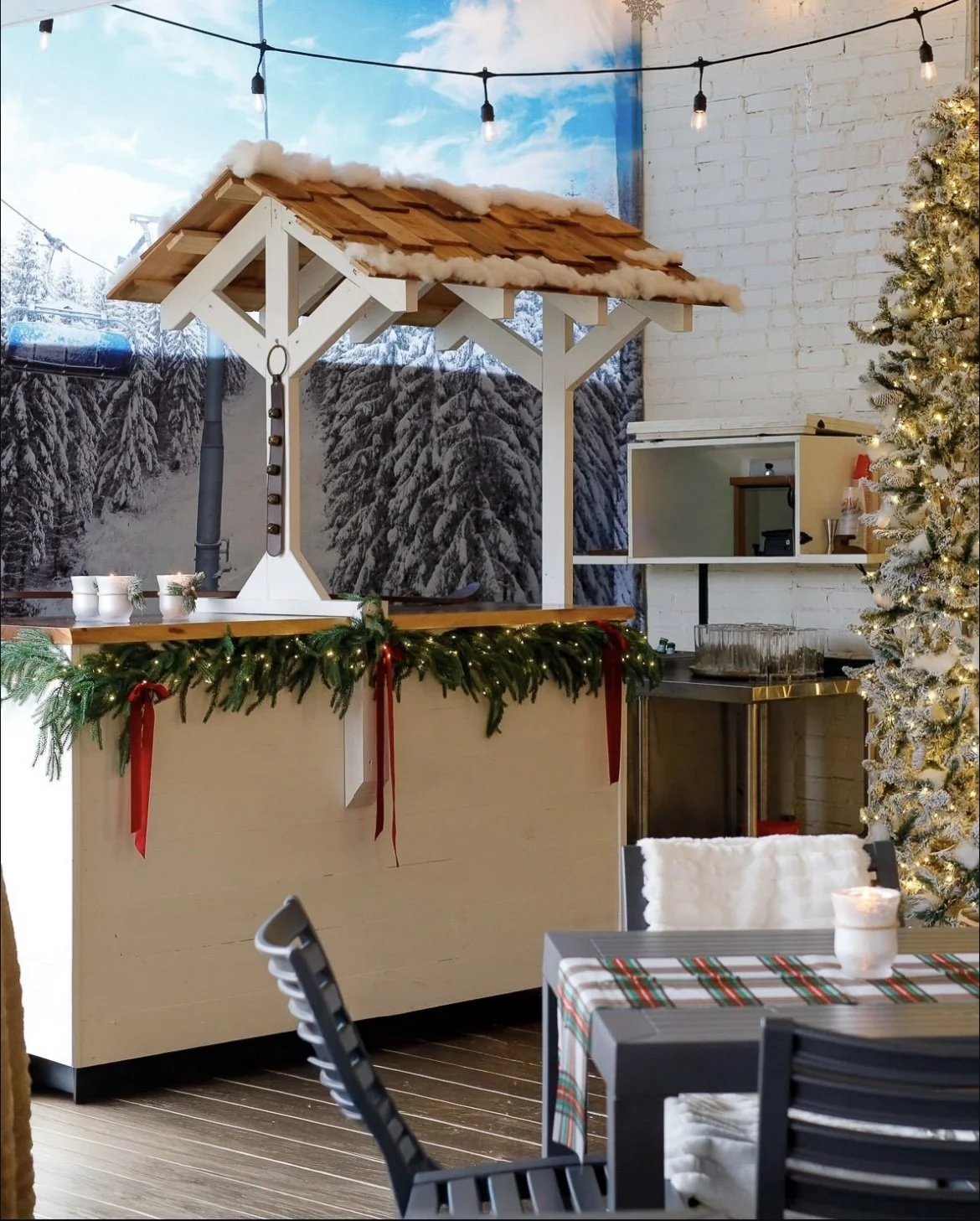 Indoor holiday food stand decorated with Christmas garland, red ribbons, and lights, with a partially constructed wooden roof structure, Christmas tree, tables, chairs, and a snowy mountain mural in the background.