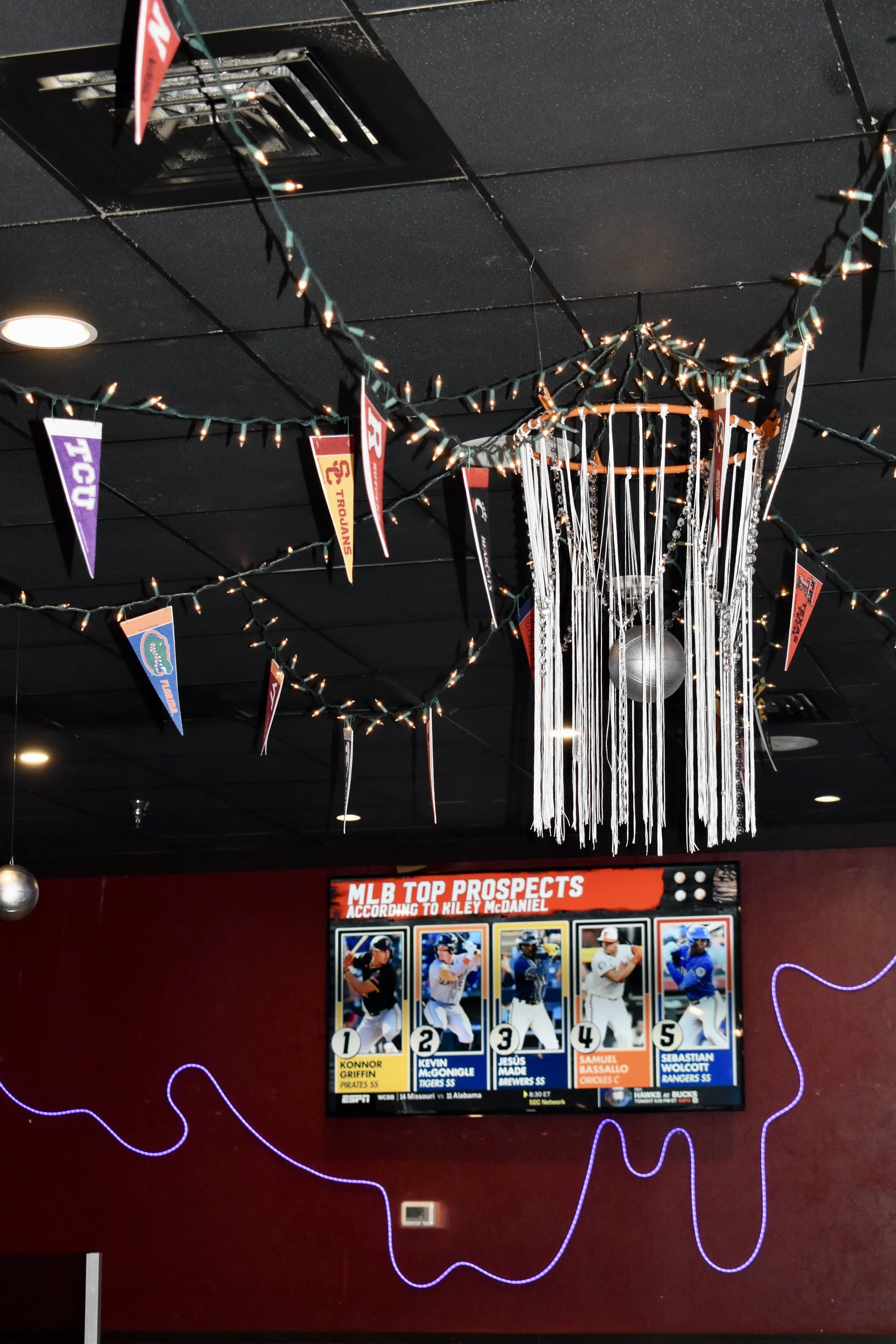 Decorated ceiling with string lights, hanging pennants of sports teams, a disco ball, and a large screen displaying top baseball prospects. March Madness