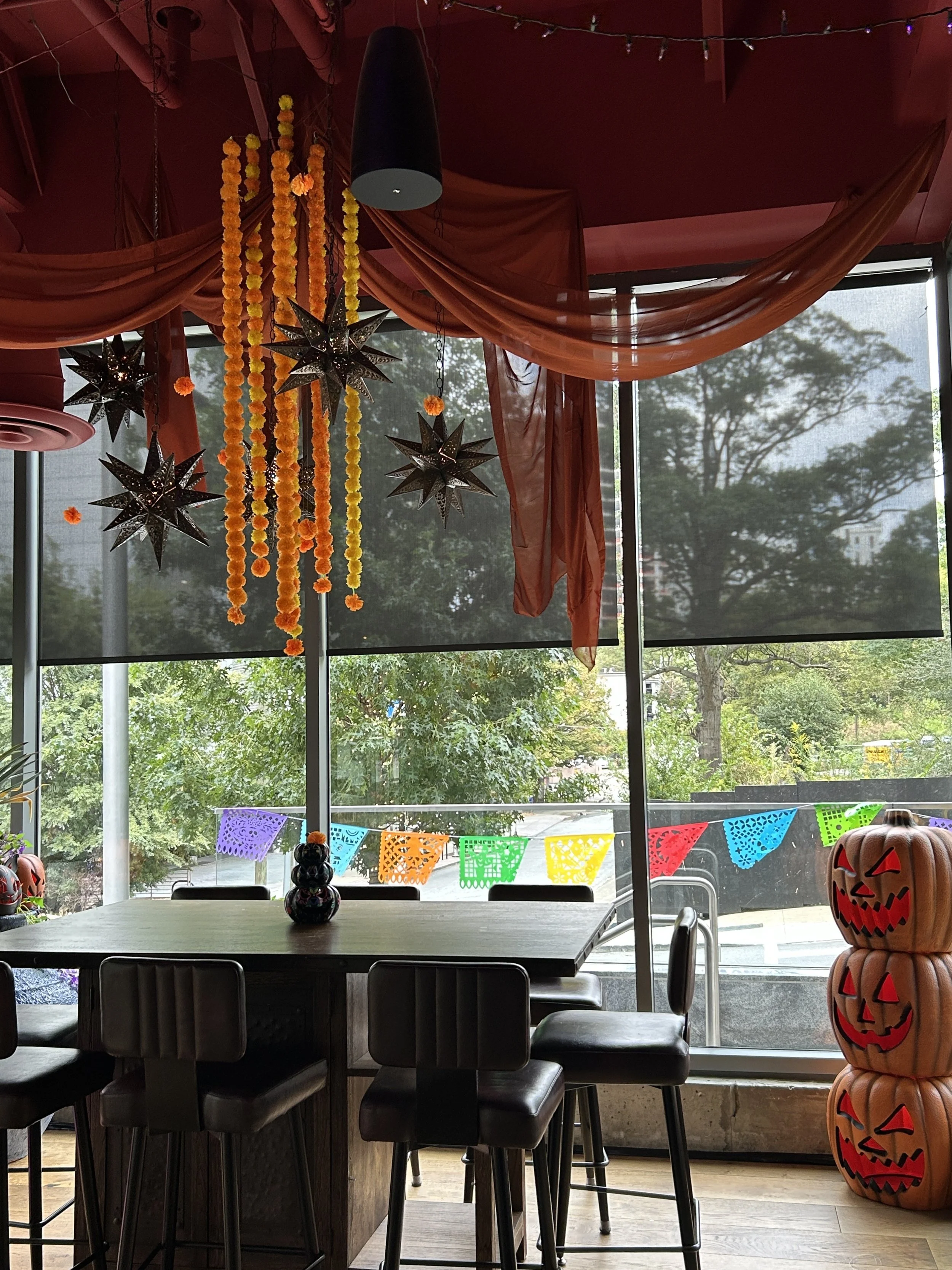 A room decorated for Halloween with paper garlands, hanging star-shaped lanterns, Halloween-themed decorations, and stacked carved pumpkins with faces, near a large window with colorful paper banners outside. Emilio's Tacos & Tequila Midtown Atlanta.