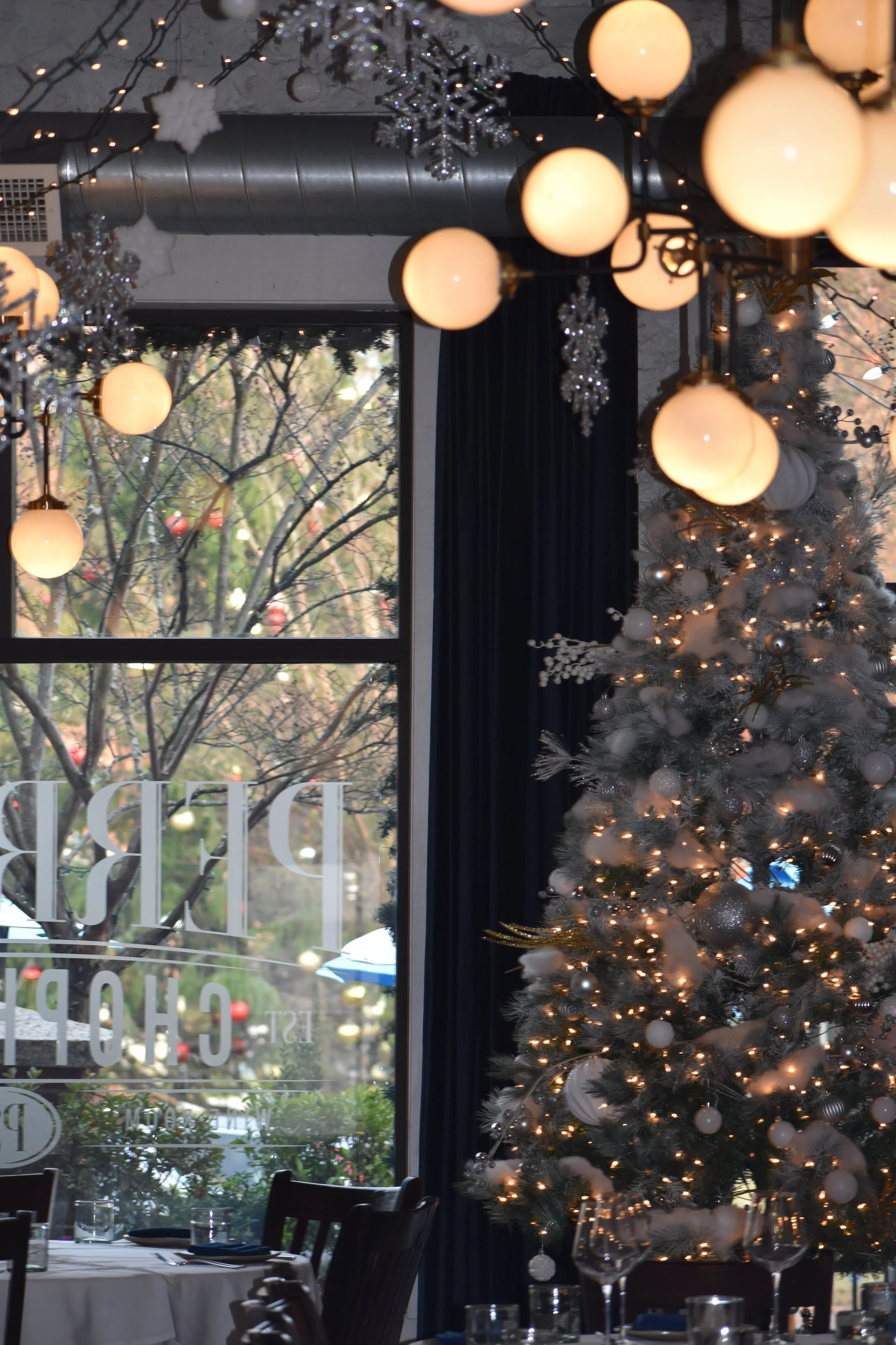 Decorated Christmas tree with white ornaments and string lights inside a restaurant, with a large window showing trees and outdoor decorations, and hanging globe lights