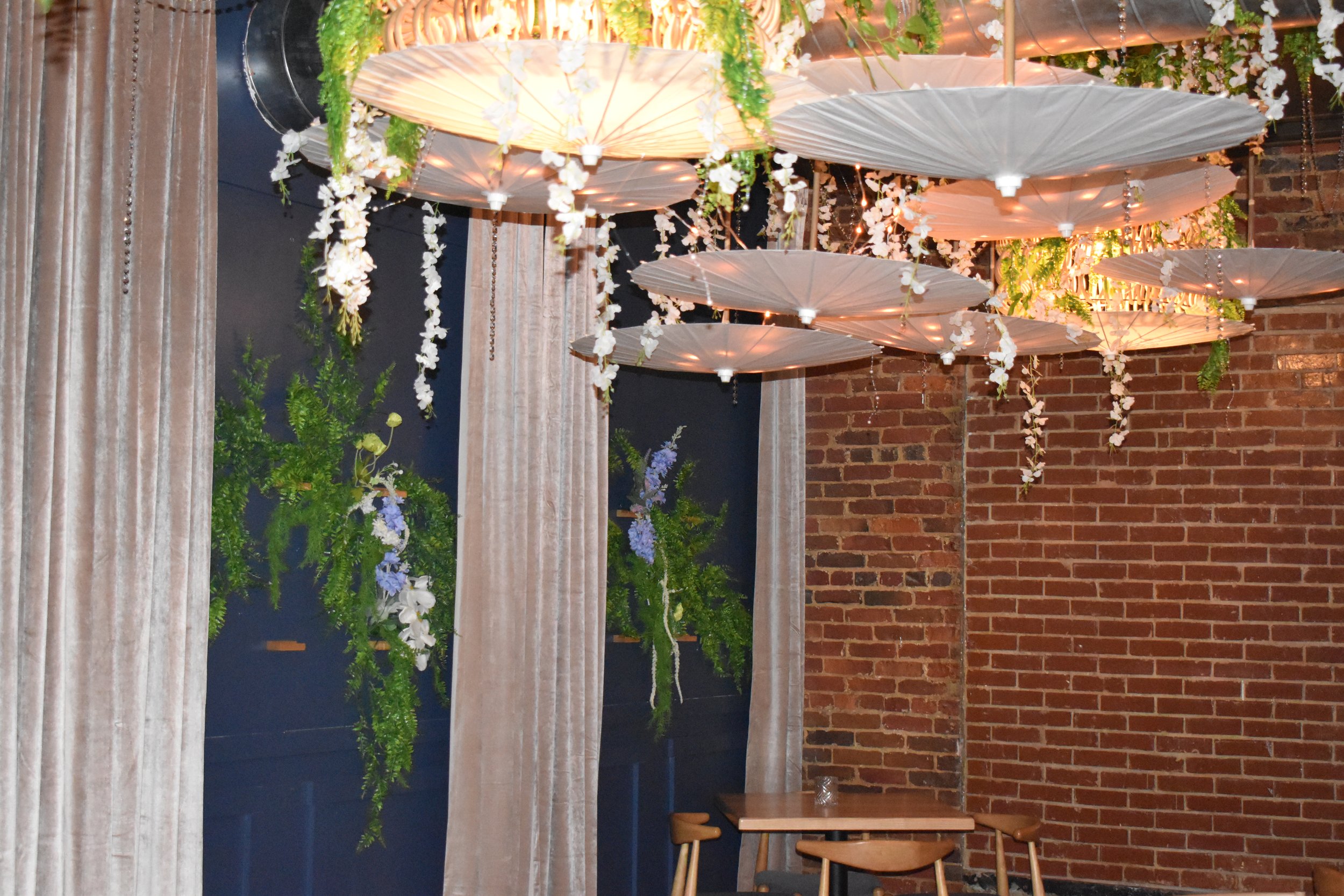 Decorated interior with hanging paper lanterns, white floral arrangements, beige curtains, a blue wall, a brick wall, small tables, and wooden chairs. Hamp & Harry's Marietta Square.