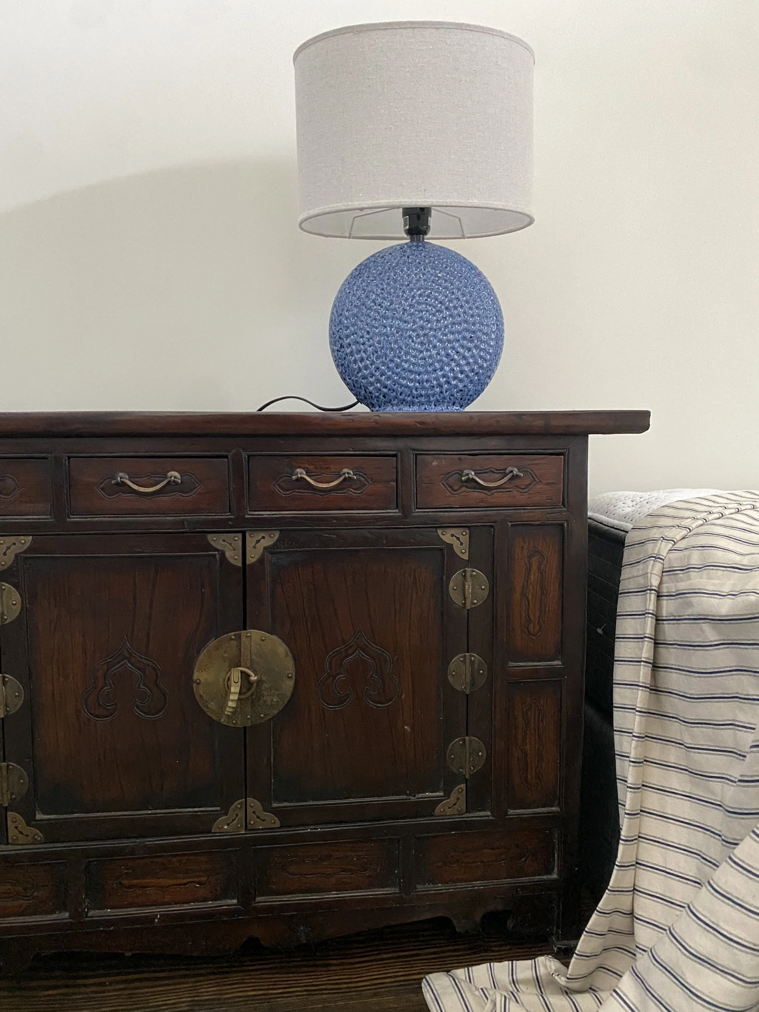 A blue textured table lamp with a white lampshade sitting on a dark wooden cabinet with ornate brass hardware, next to a bed with striped bedding. Hilton Head Island Interior Design.