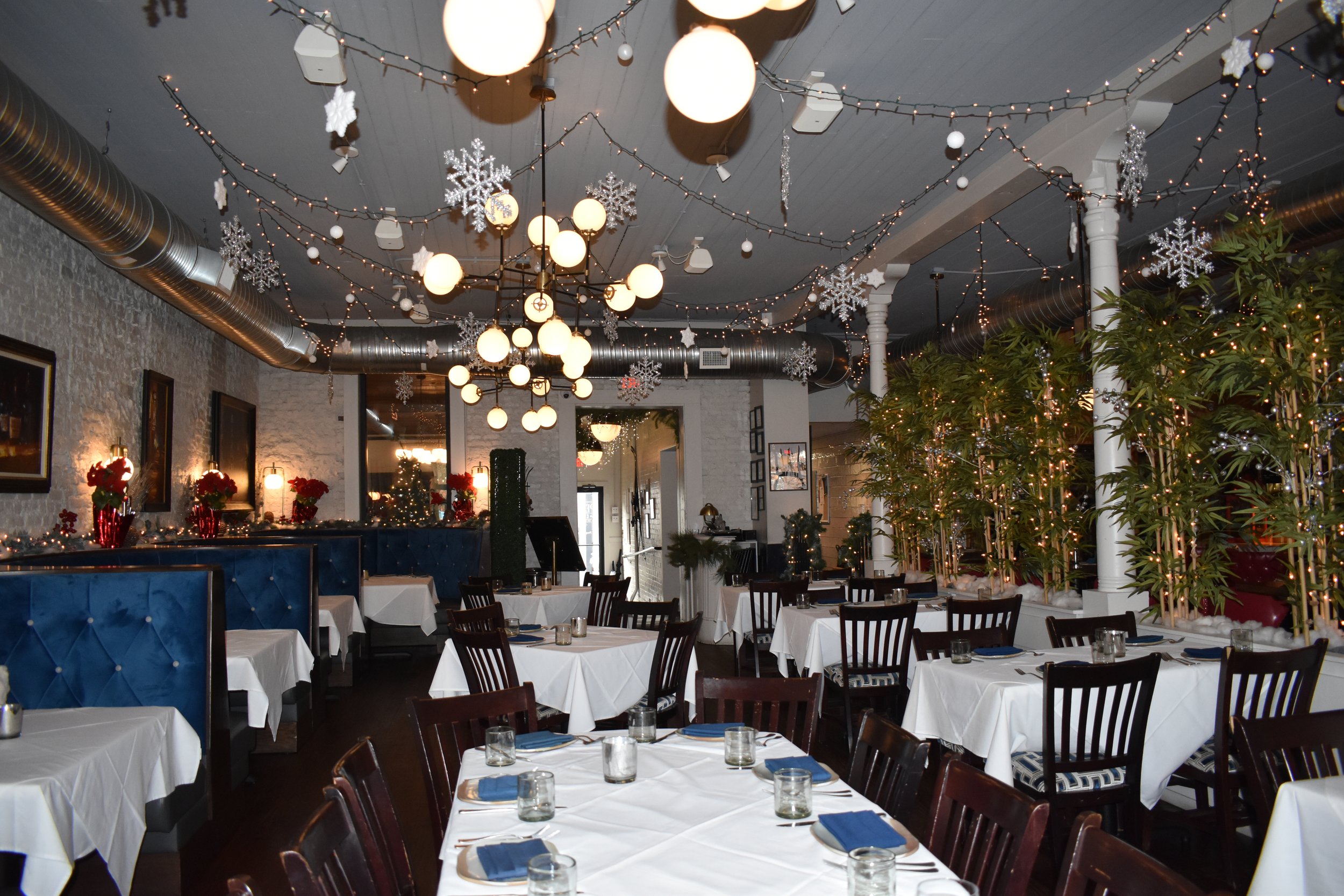 A decorated restaurant with white tablecloths, blue napkins, glasses, and cutlery, featuring holiday decorations including snowflakes, Christmas trees, and string lights. Perry St Chophouse.