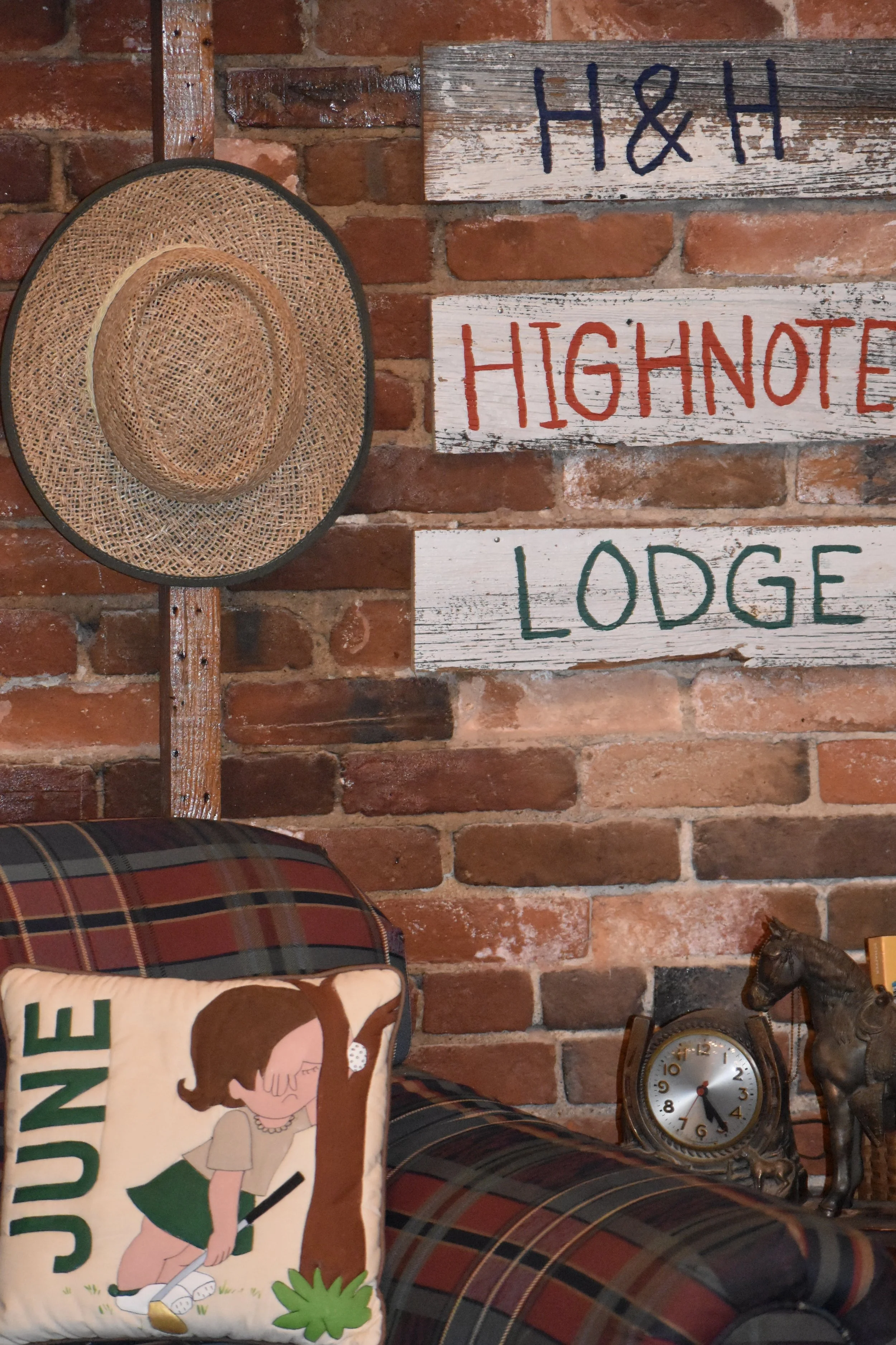 Brick wall with decorative signs reading "H&H," "High Note," and "Lodge," a woven hat hanging on a wooden hook, a plaid couch with an illustrated cushion with the word "June," a vintage clock, and a brass horse figurine.