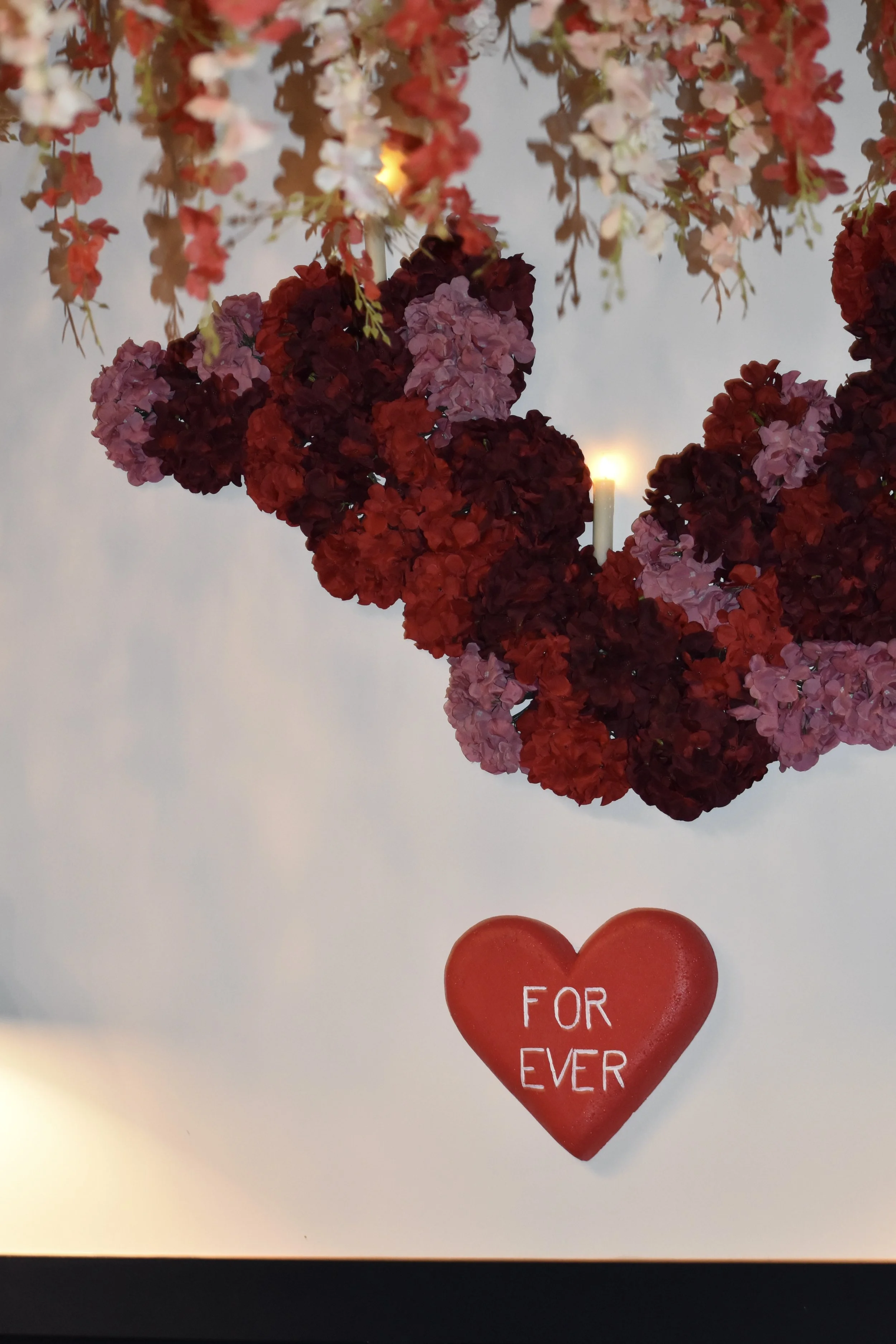 A heart-shaped decoration with the words "FOR EVER" written on it, hanging from a floral arrangement with pink, red, and purple flowers.