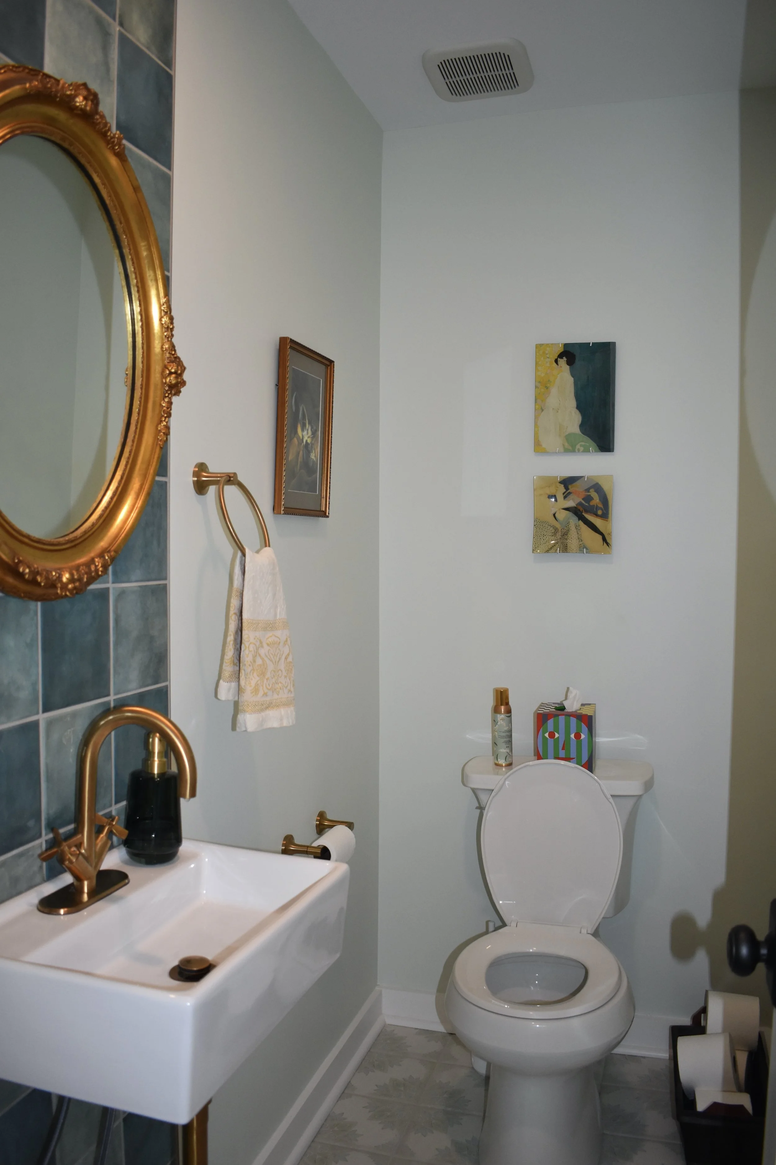 A small bathroom with blue tiled walls and a white porcelain sink with a gold faucet. A small mirror with a gold frame is above the sink, and a hand towel hangs from a gold towel ring. Across from the sink is a white toilet with a container of spray 