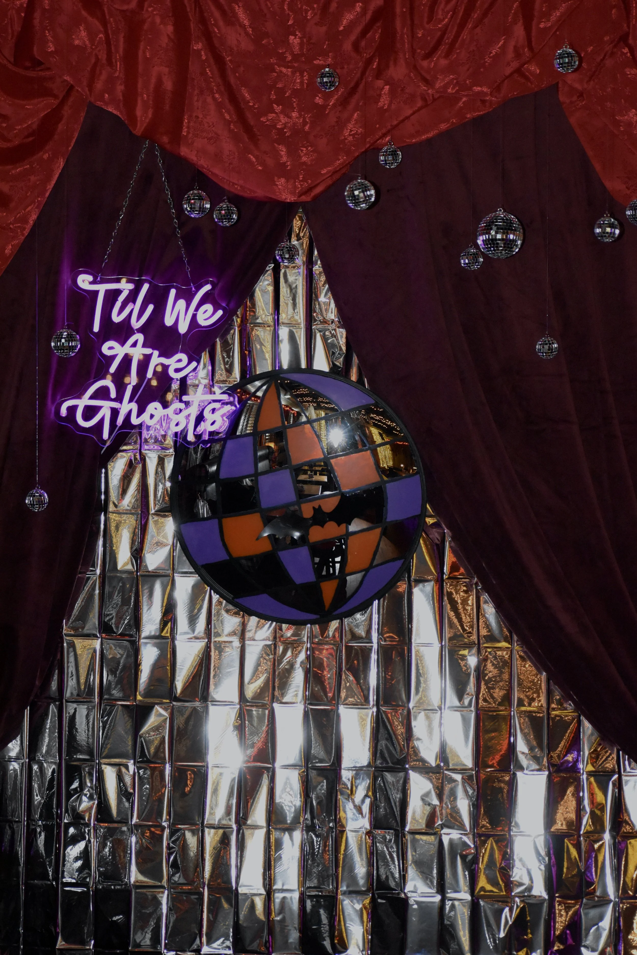 Decorative background with red velvet curtains, hanging mirrored disco balls, a neon sign that reads 'Til We Are Ghosts,' and a multicolored globe with a black bat silhouette inside.