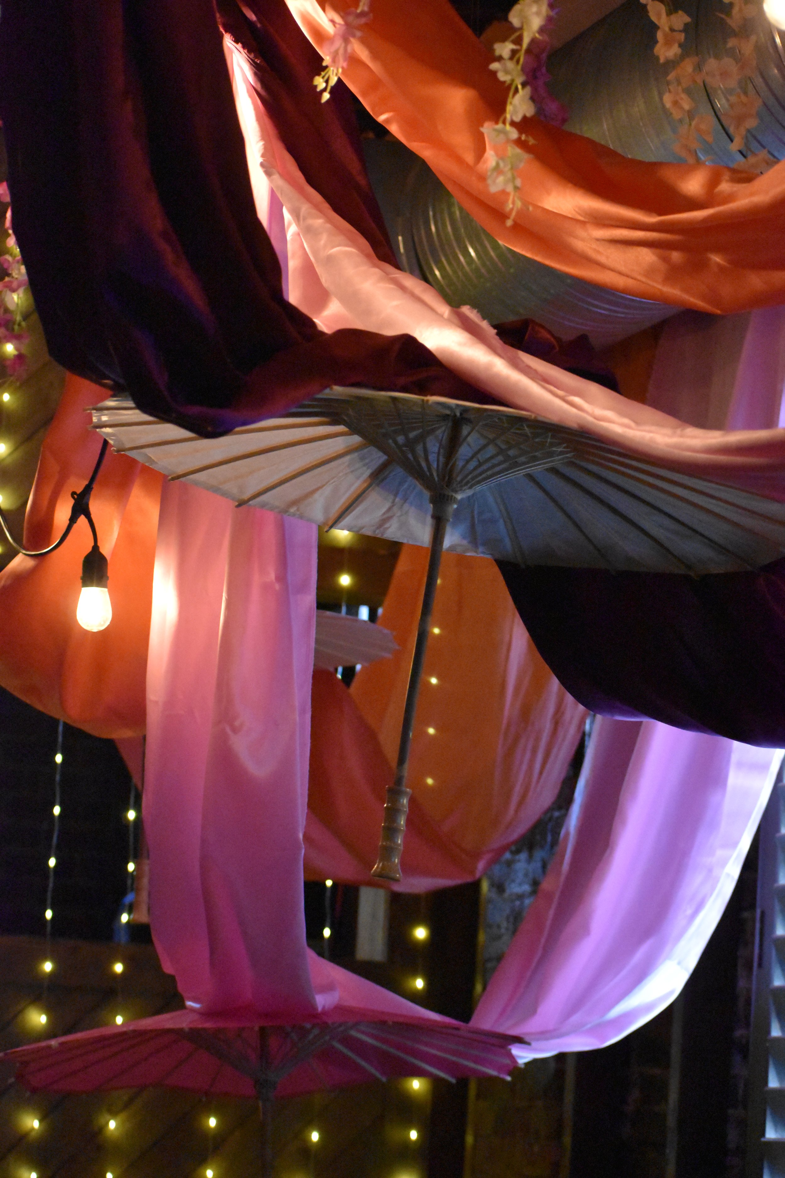 Decorative setup with pink, purple, and orange fabric drapes, umbrellas hanging upside down, and string lights on a dark background. Hamp & Harry's Marietta Square.