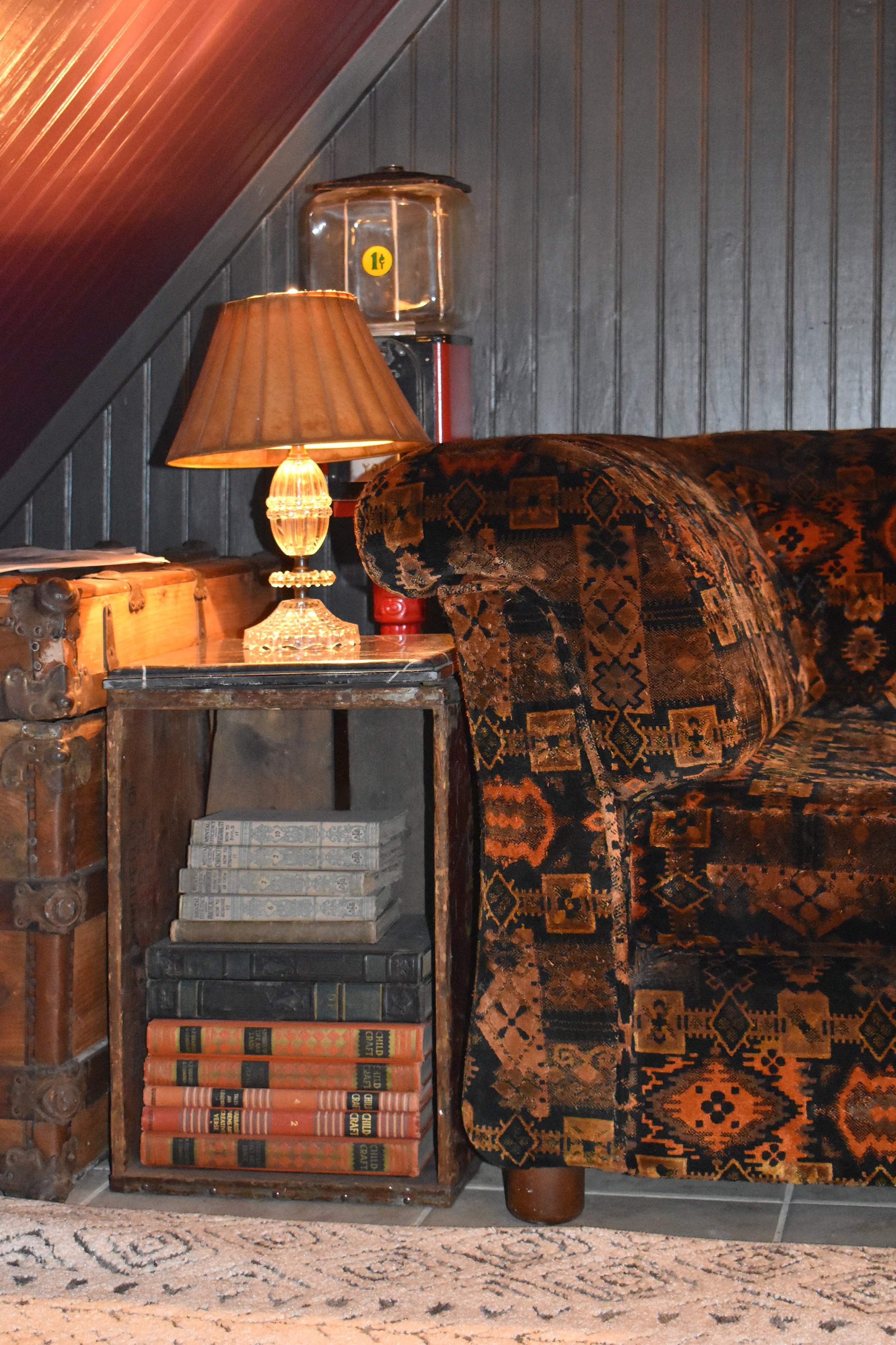 A cozy corner with a vintage patterned armchair, a wooden side table with a glass lamp, and a stack of old books in a room with wood-paneled walls.