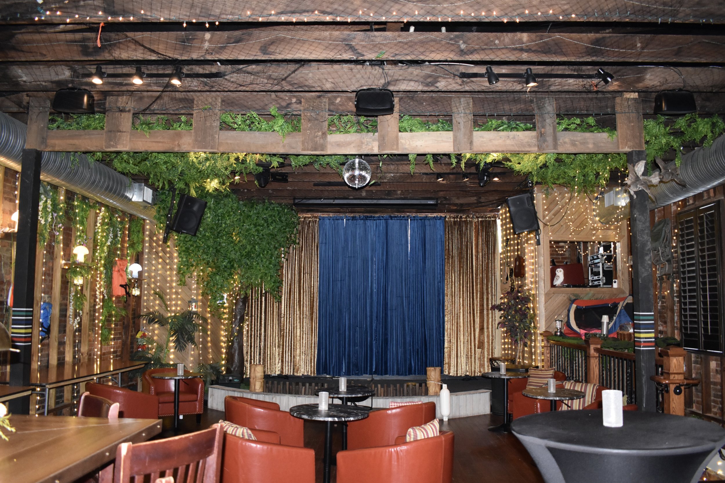 Interior of a decorated event space or restaurant with a stage, blue curtain, fairy lights, greenery, wooden beams, and seating. Hamp & Harry's Marietta Square.