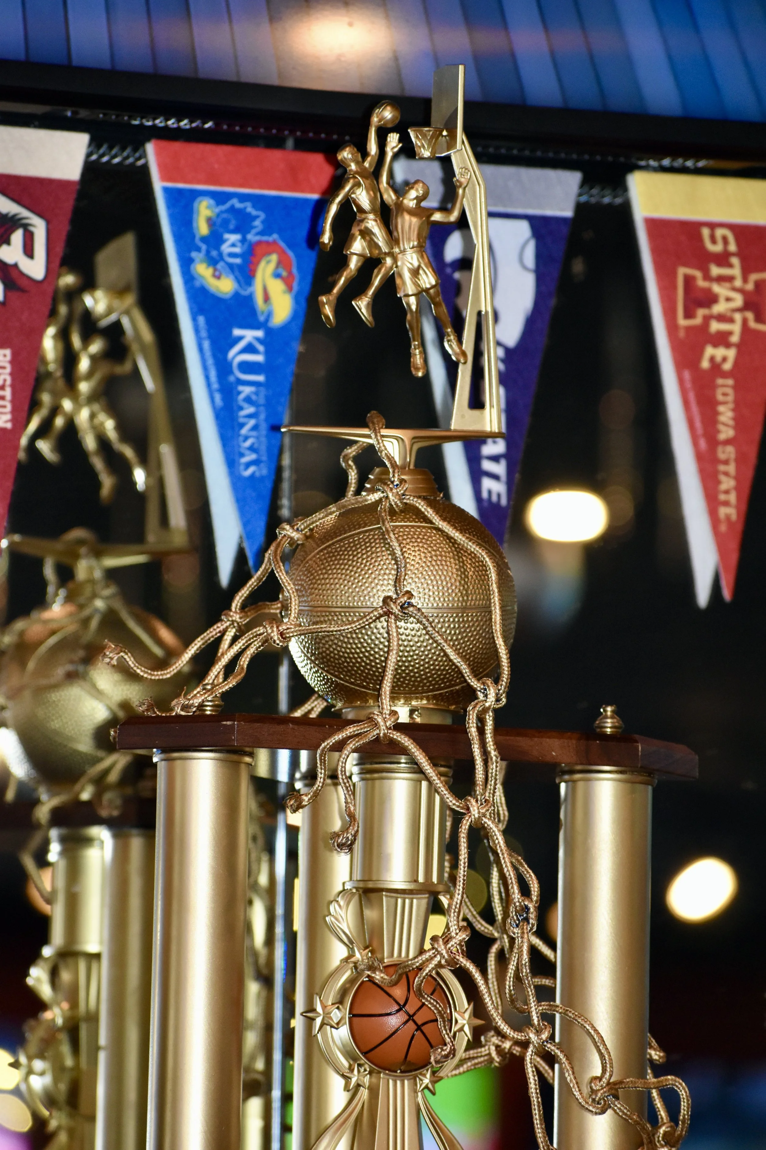Close-up of a basketball trophy with figures of basketball players, basketballs, and gold-colored columns, with Kansas and Missouri Royals banners in the background. March Madness