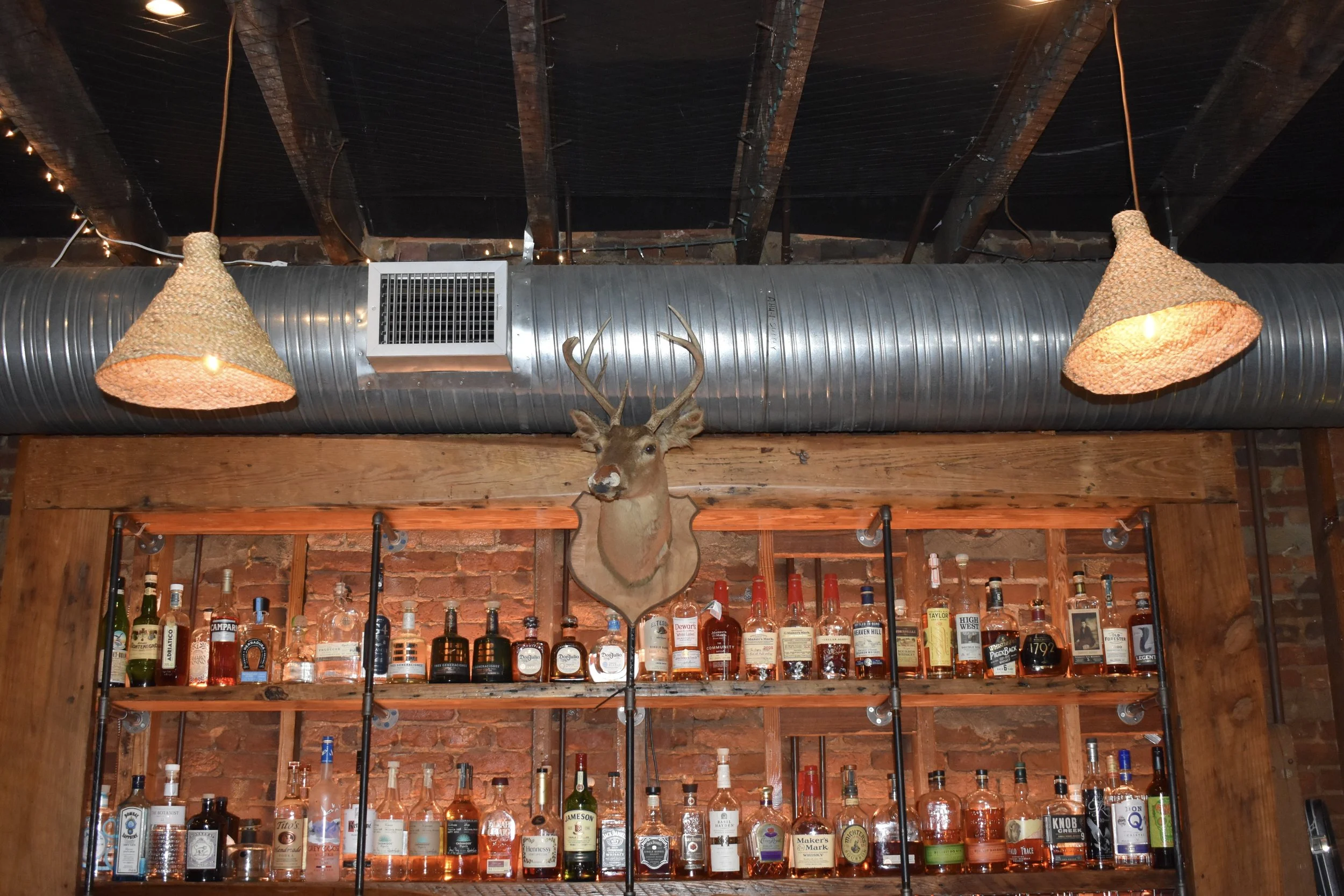 A bar with shelves of various bottles of liquor mounted on a brick wall, a mounted deer head with antlers on the wall above, and two woven pendant lights hanging from the ceiling.