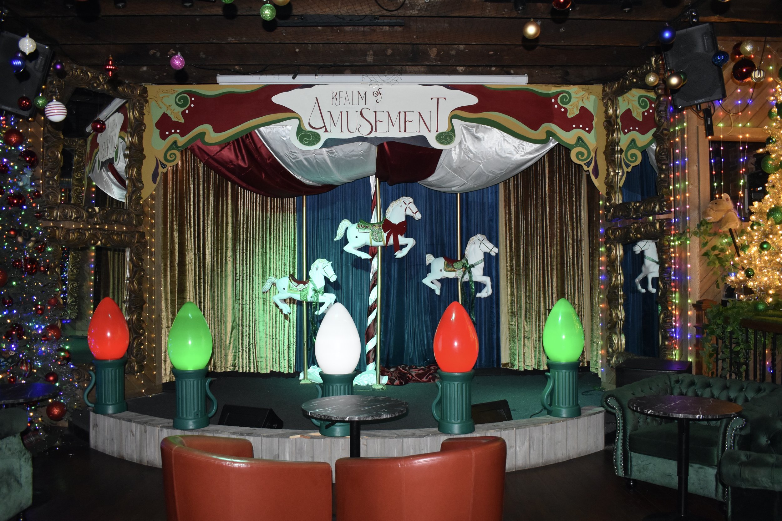 A small stage decorated with a carousel theme, featuring plush carousel horses, Christmas ornaments, and colorful lighting. The backdrop has a sign that reads 'Realm of Amusement' and gold curtains, with surrounding Christmas trees decorated with lig