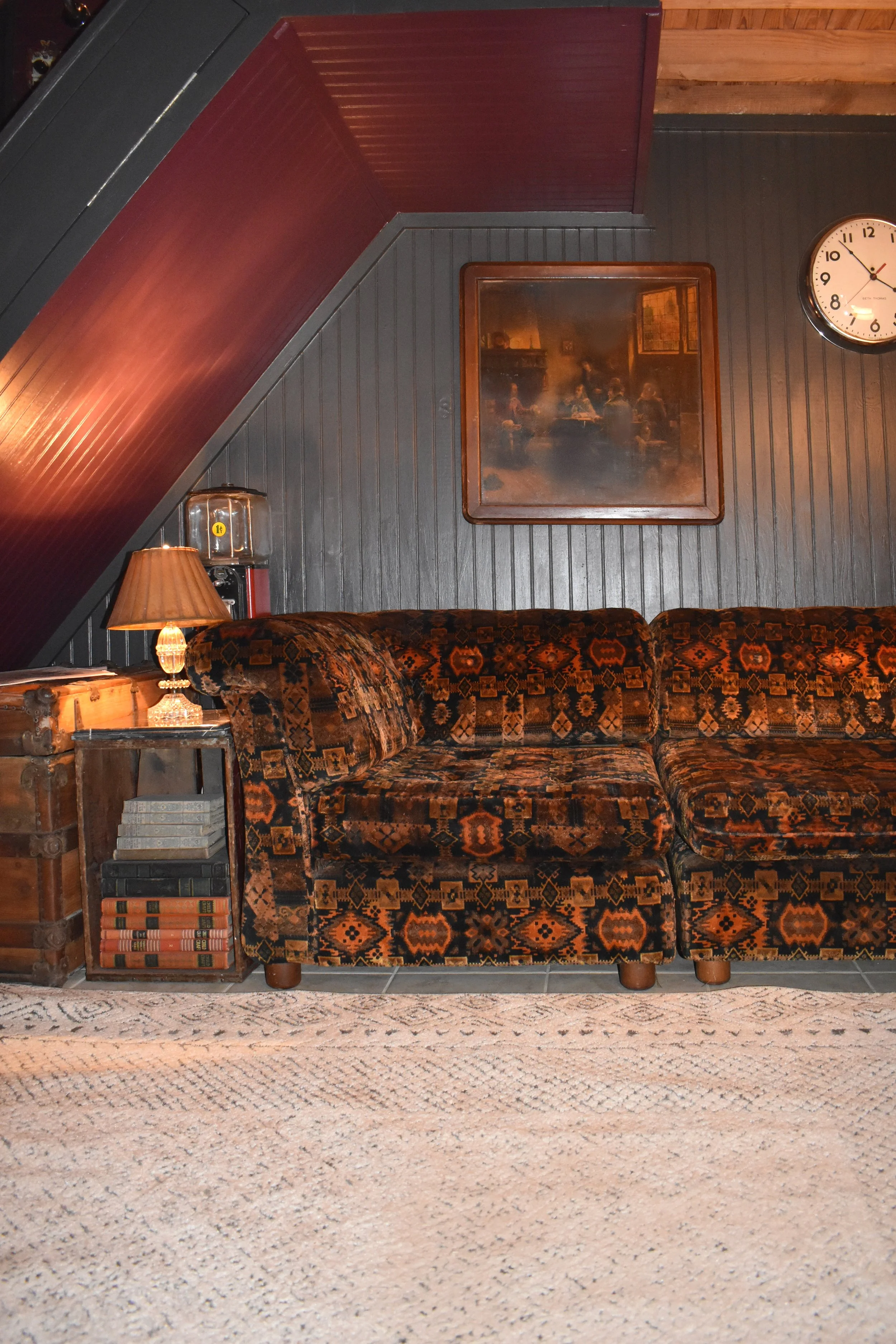 A vintage-style room with a patterned sofa, a wooden side table with books, a lamp, a wall clock, and framed artwork on wooden-paneled walls.