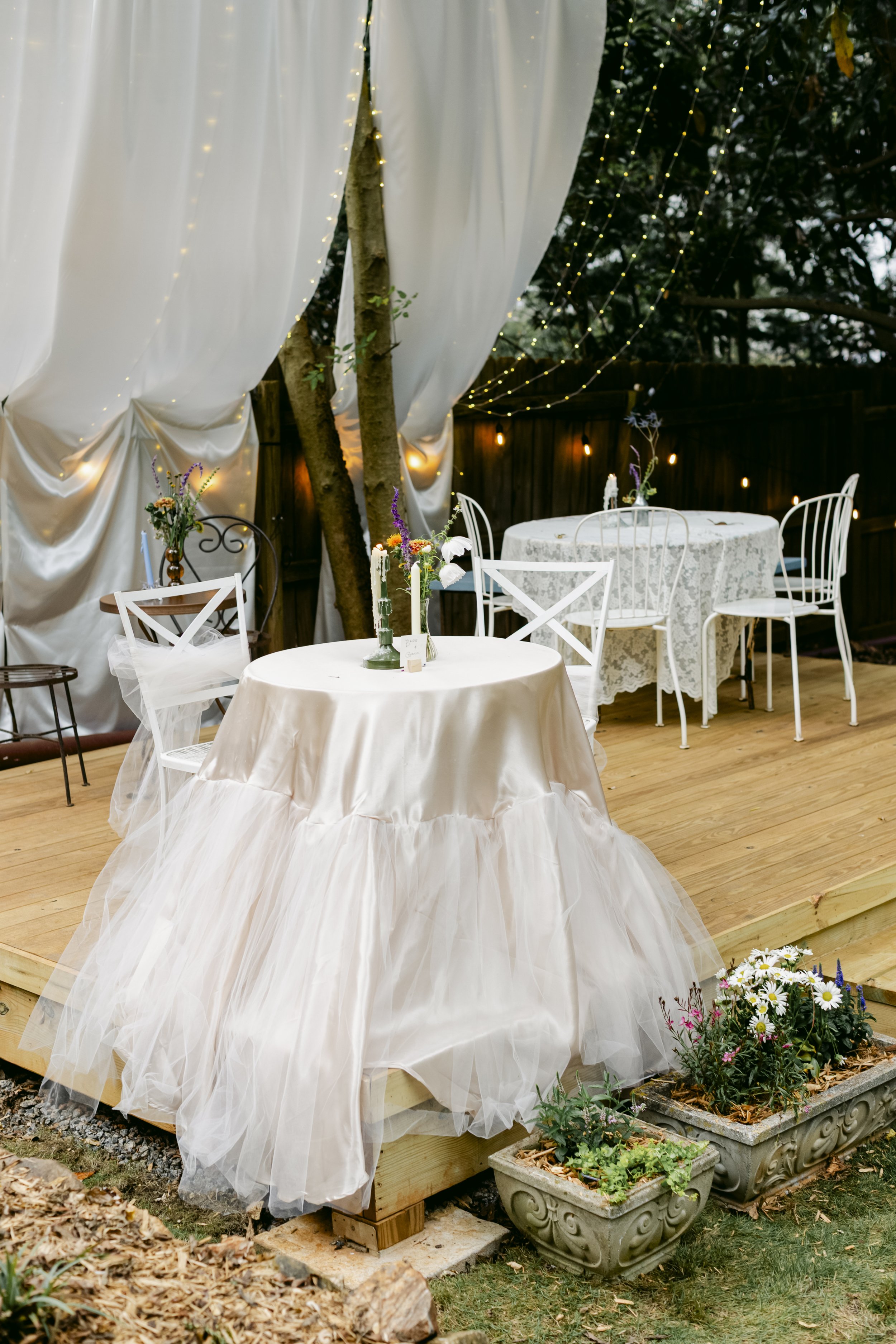 Outdoor wedding reception setup on a wooden deck with round tables covered in lace and satin tablecloths, surrounded by white chairs, flowers, and string lights. Whimsical backyard wedding with wildflowers, fun colors, and timeless elegance. 