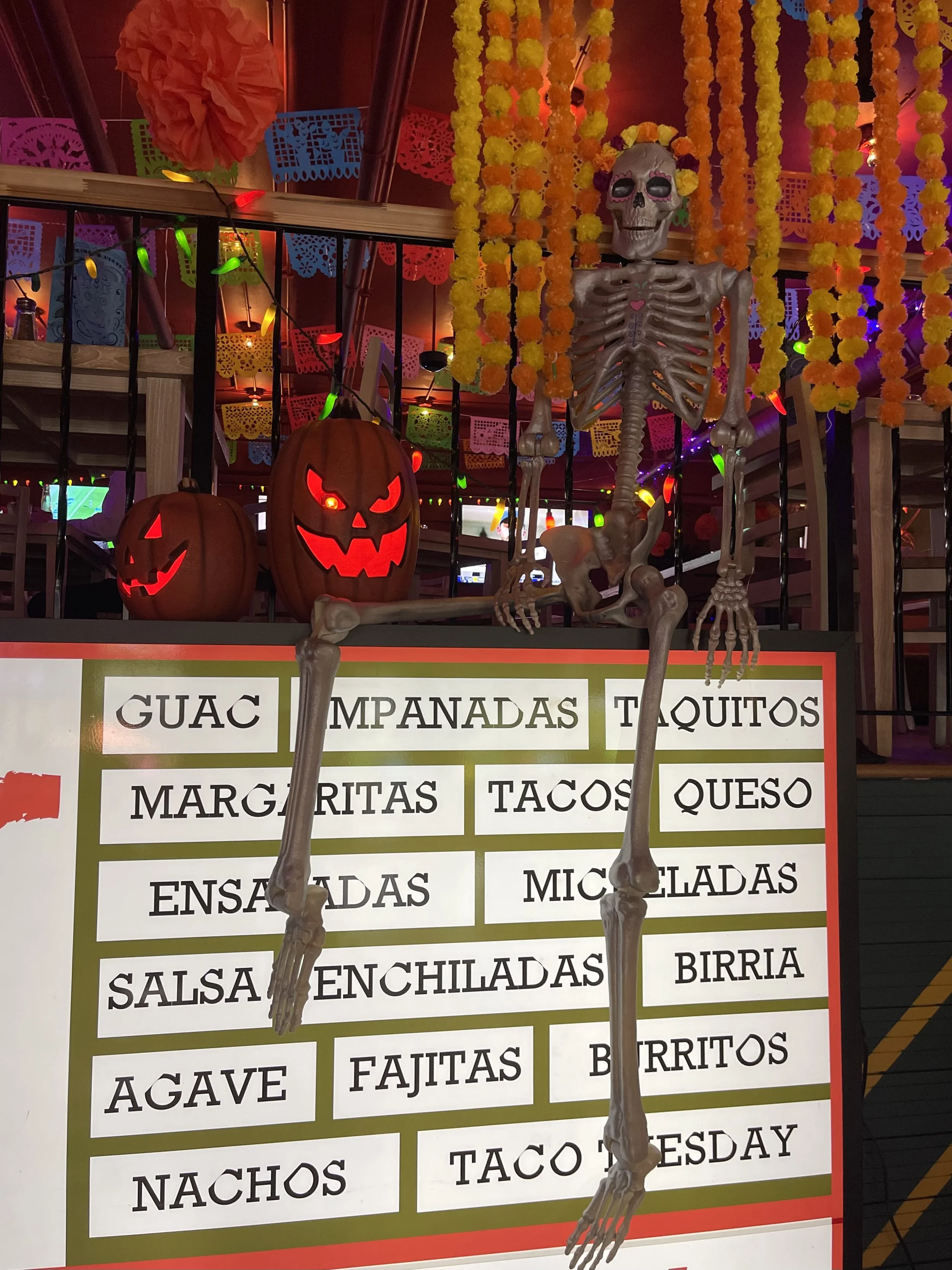 Decorative Halloween scene featuring a skeleton hanging from the ceiling, illuminated Jack-o'-lanterns, colorful papel picado banners, and a menu board with Mexican food options. Emilio's Tacos & Tequila Midtown Atlanta.