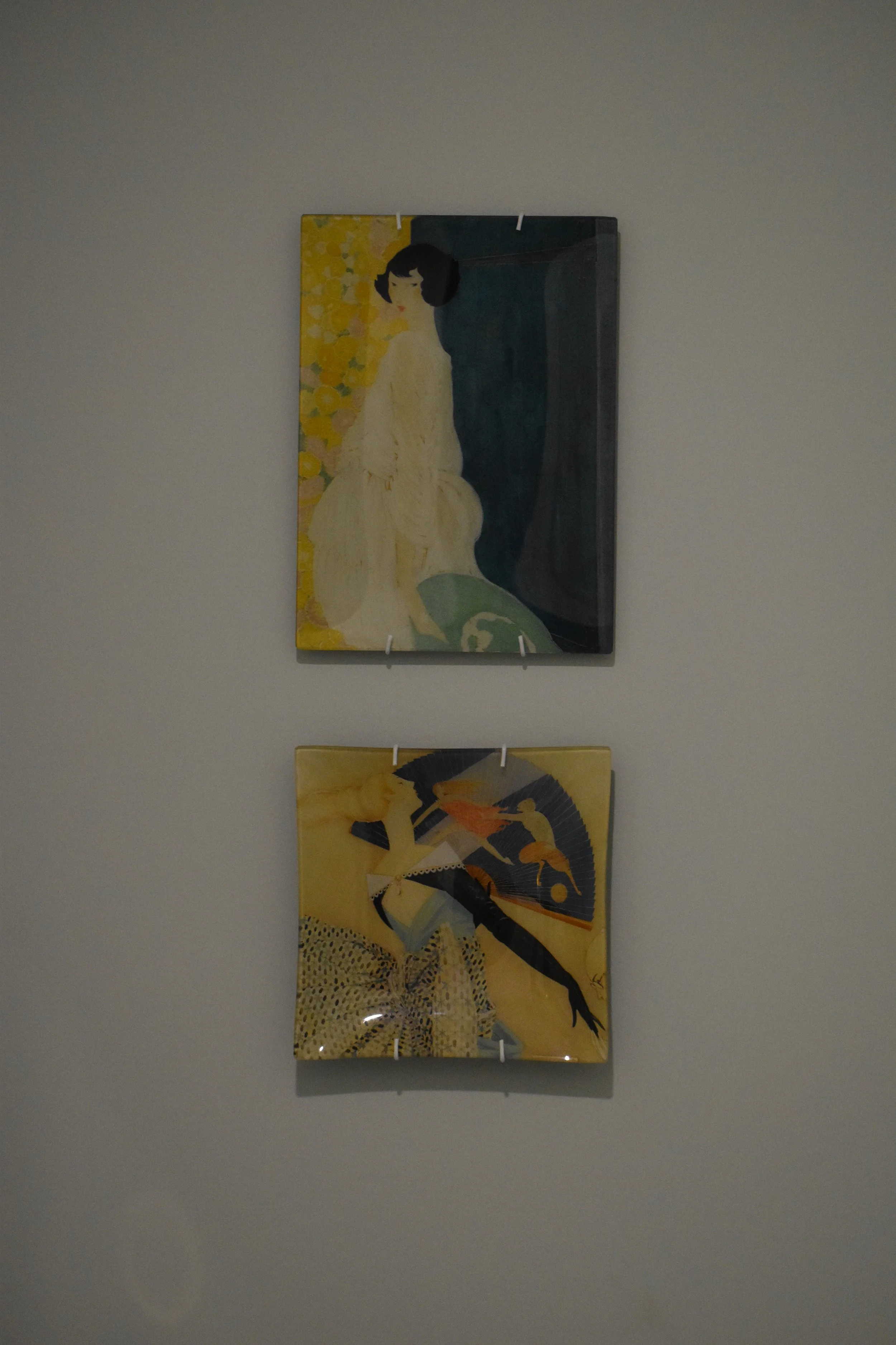 Two paintings hanging vertically on a white wall. The top painting depicts a woman with dark hair in a cream-colored dress, sitting next to a dark curtain and yellow flowers. The bottom painting shows a woman holding a parasol, dressed in a light-col