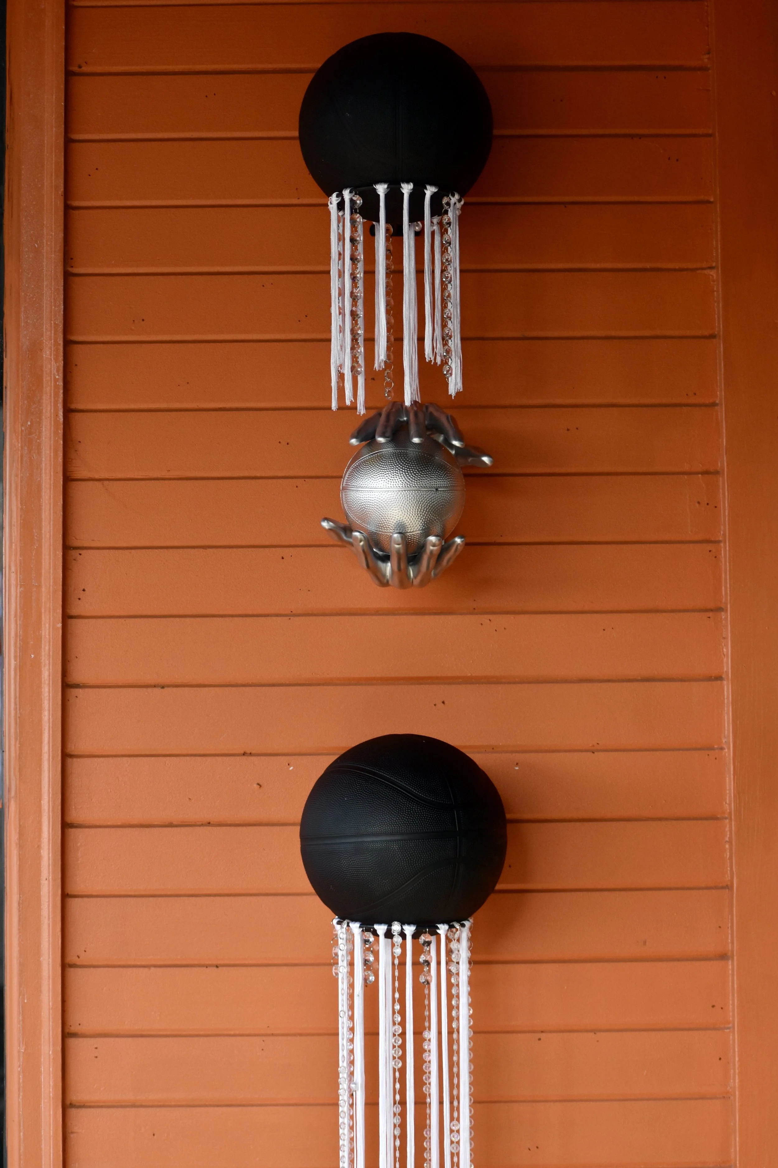 Decorative wall art with two black basketballs and a silver basketball with hand sculptures, hanging on an orange wooden wall. March Madness