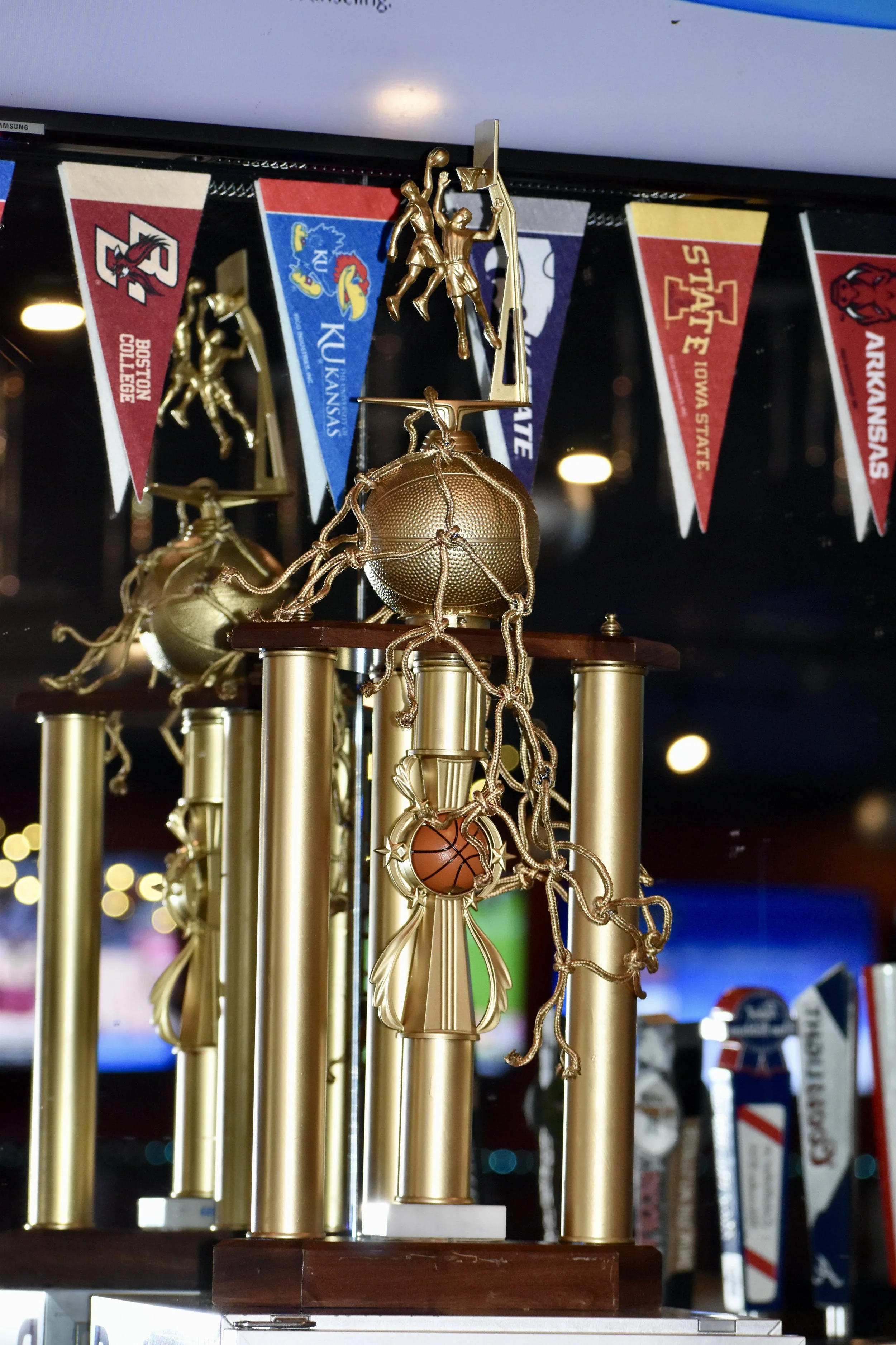A sports trophy featuring a basketball player climbing a pole, with a basketball at the top. The trophy is gold-colored with multiple columns and has basketball-themed decorations, including a small basketball pendant. It is displayed with college pe