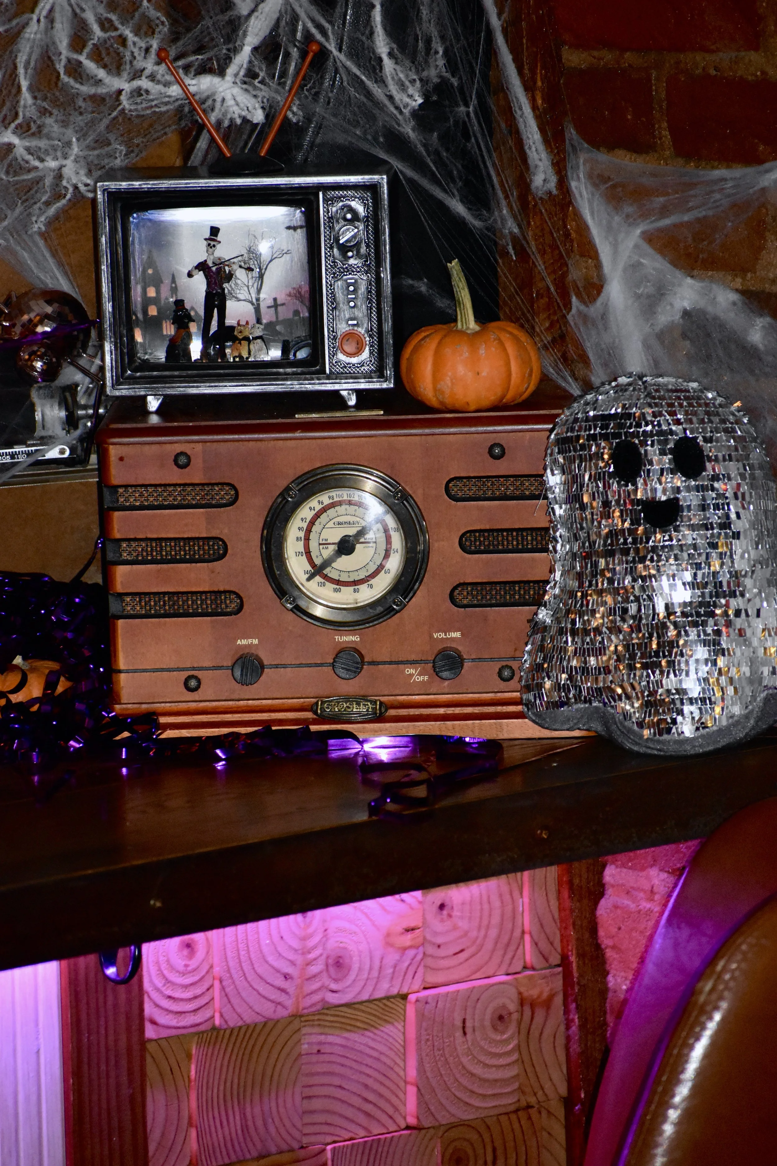 Decorative Halloween display featuring a vintage radio, a silver reflective ghost figure, a small pumpkin, a mini television with a skeleton figurine playing a violin on screen, and spider web decorations.