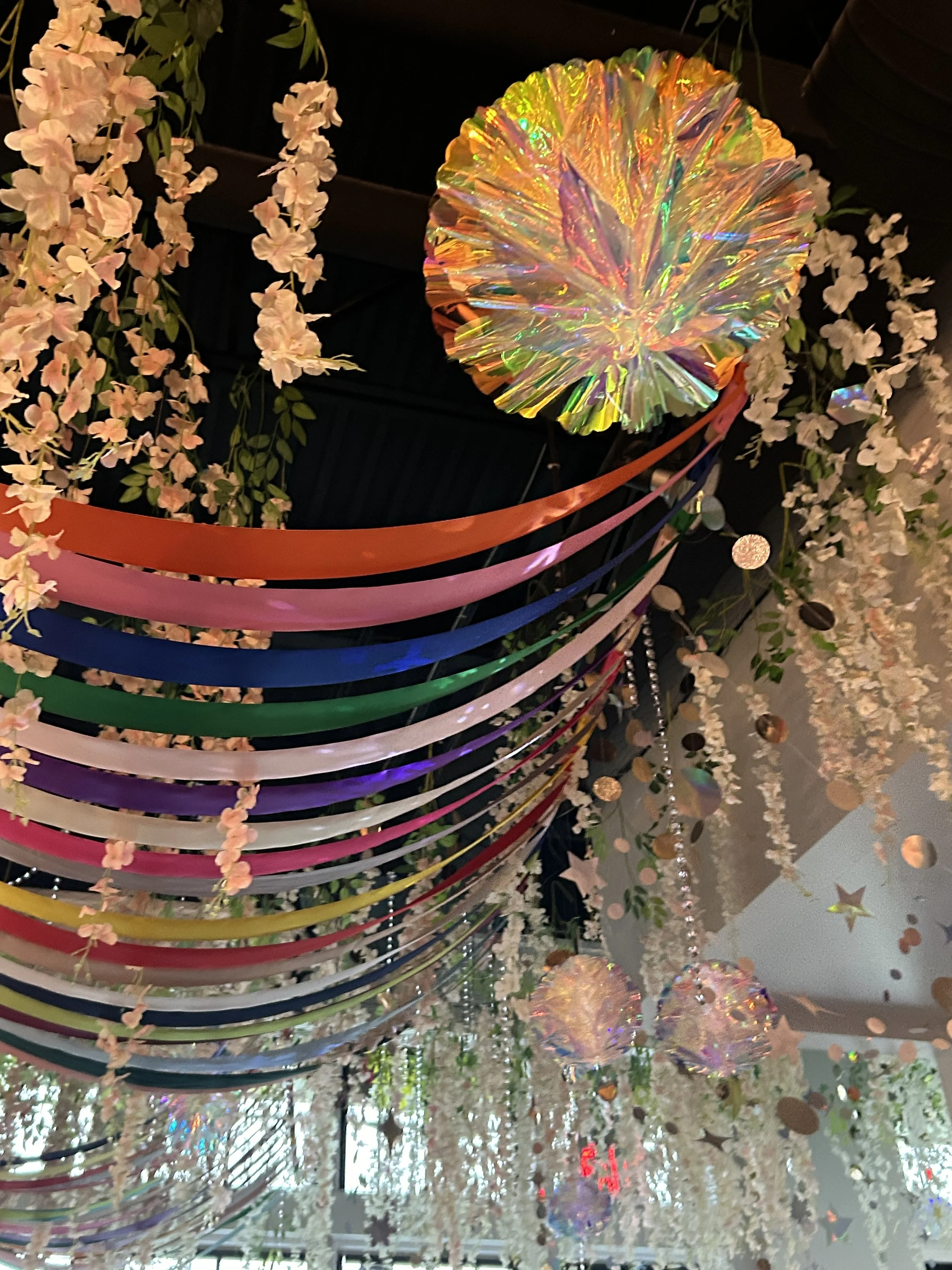 Colorful iridescent paper decorations hanging from the ceiling with artificial flowers and stars.