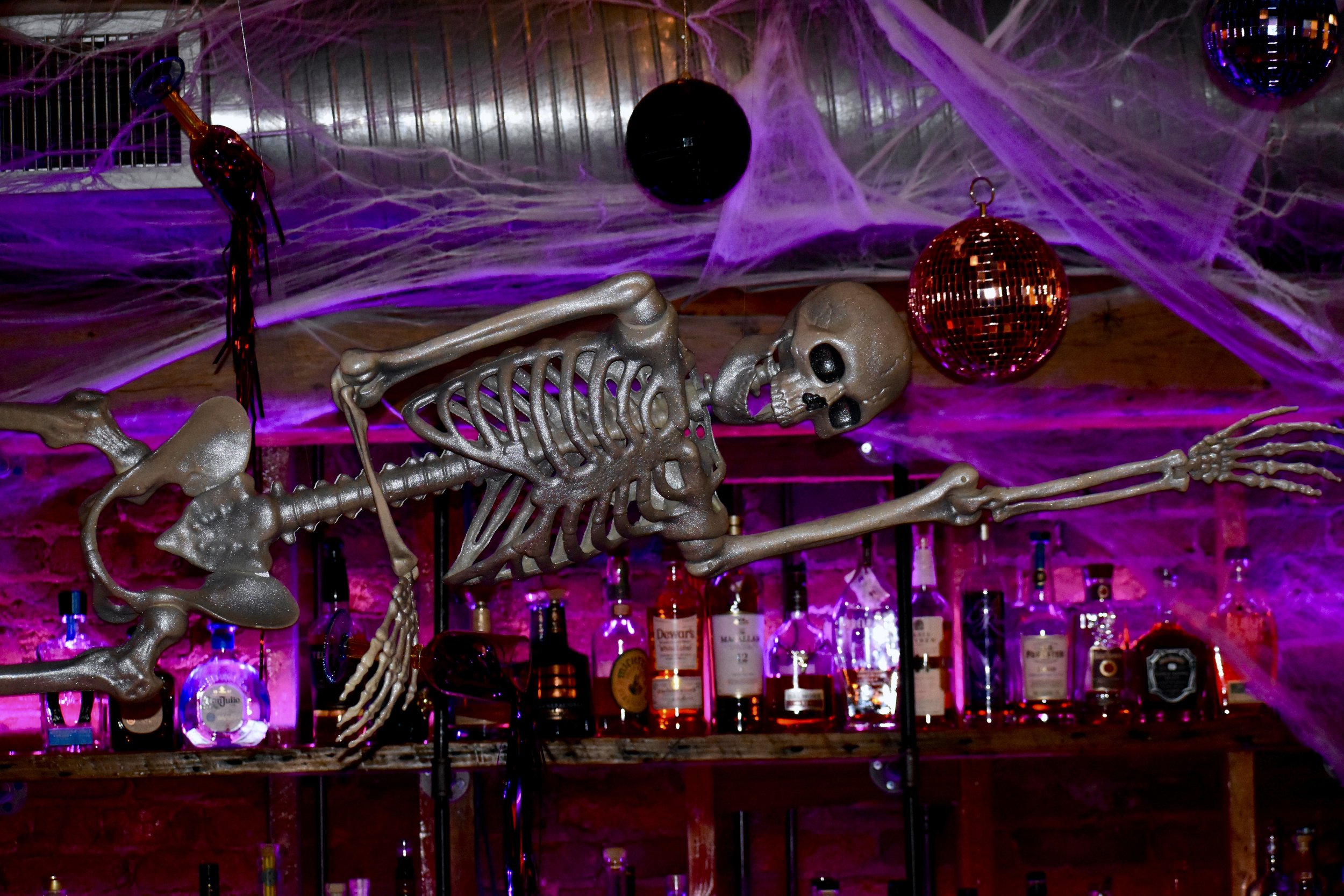 Decorated Halloween bar with a skeleton figure hanging from the ceiling, spider webs, and Halloween ornaments, illuminated by purple lighting.
