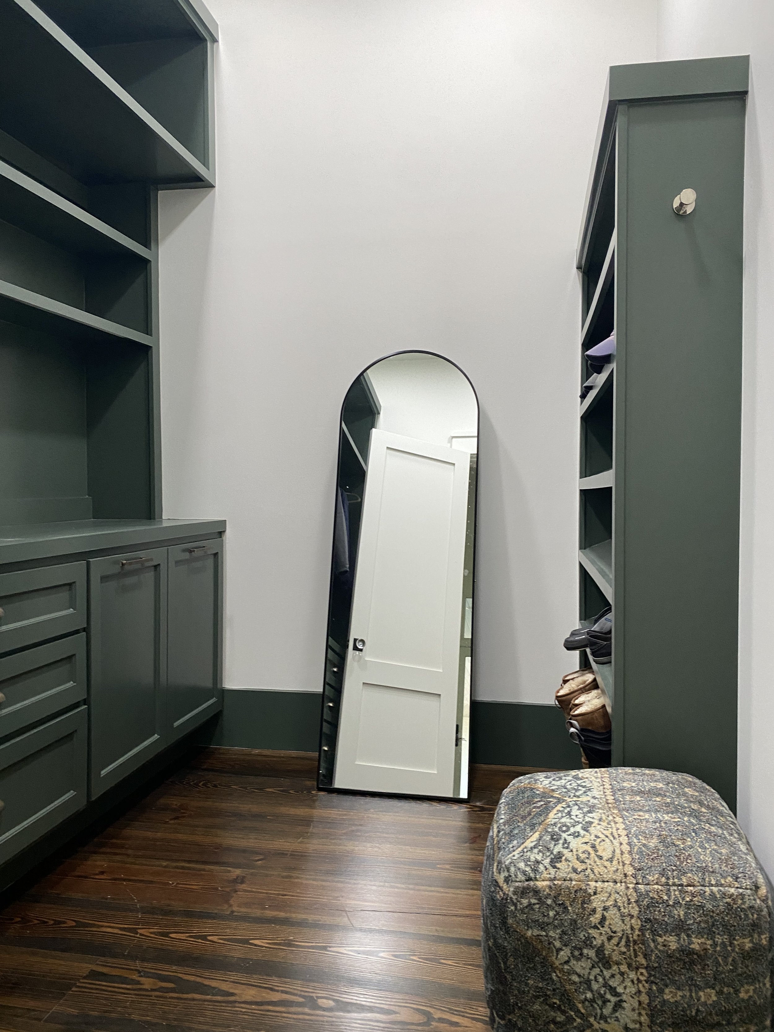A room with green built-in shelves and cabinets, a tall mirror leaning against the wall, and a small upholstered footstool with a patterned fabric. The room has wood flooring and neutral wall colors. Hilton Head Island Interior Design.