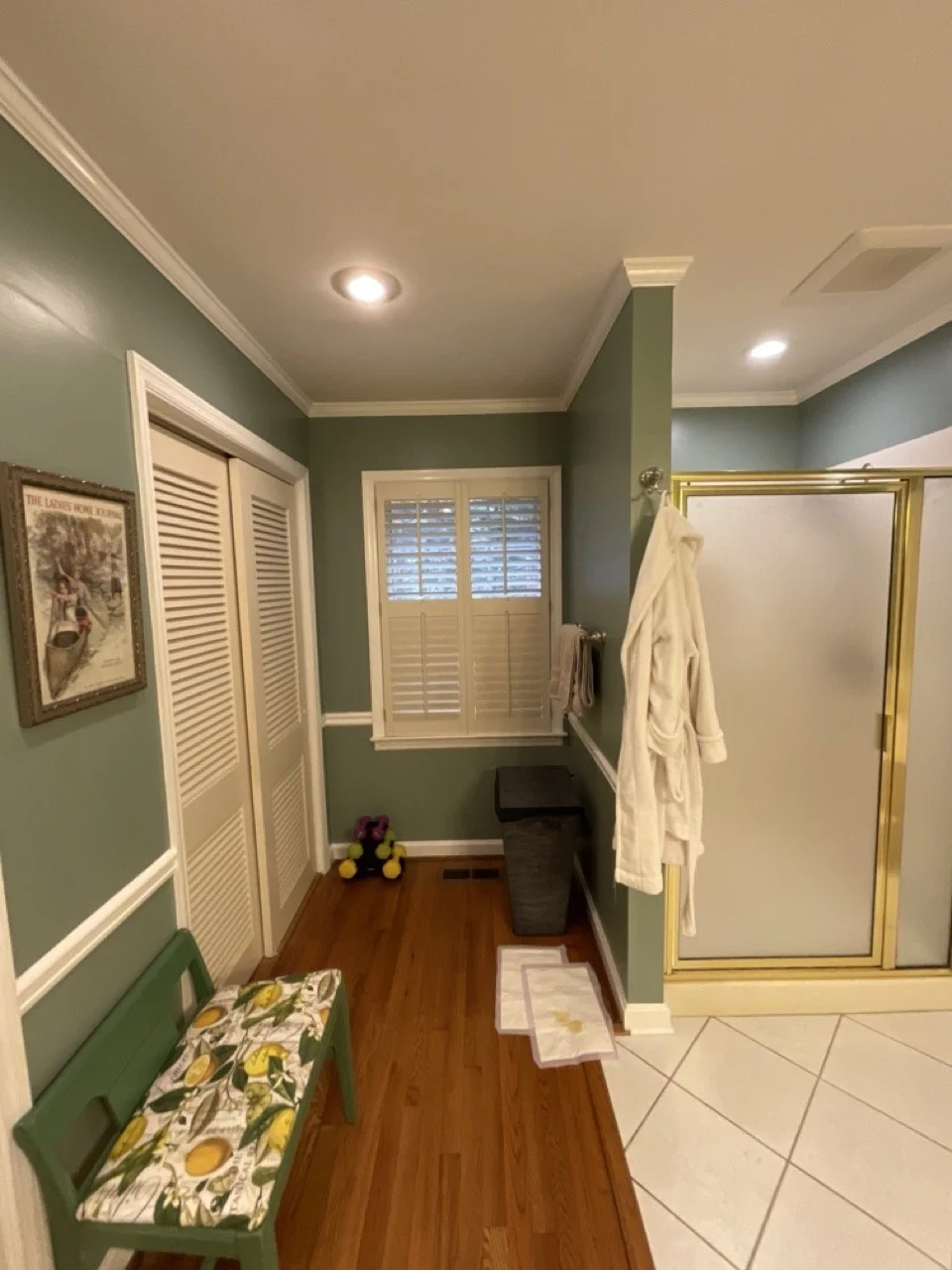 Bathroom with hardwood floor, green walls, and white crown molding. Features a green bench with lemon print cushion, a framed picture on the wall, a window with white shutters, a laundry hamper, a small pair of dumbbells, white robe hanging, and a shower with gold trim.