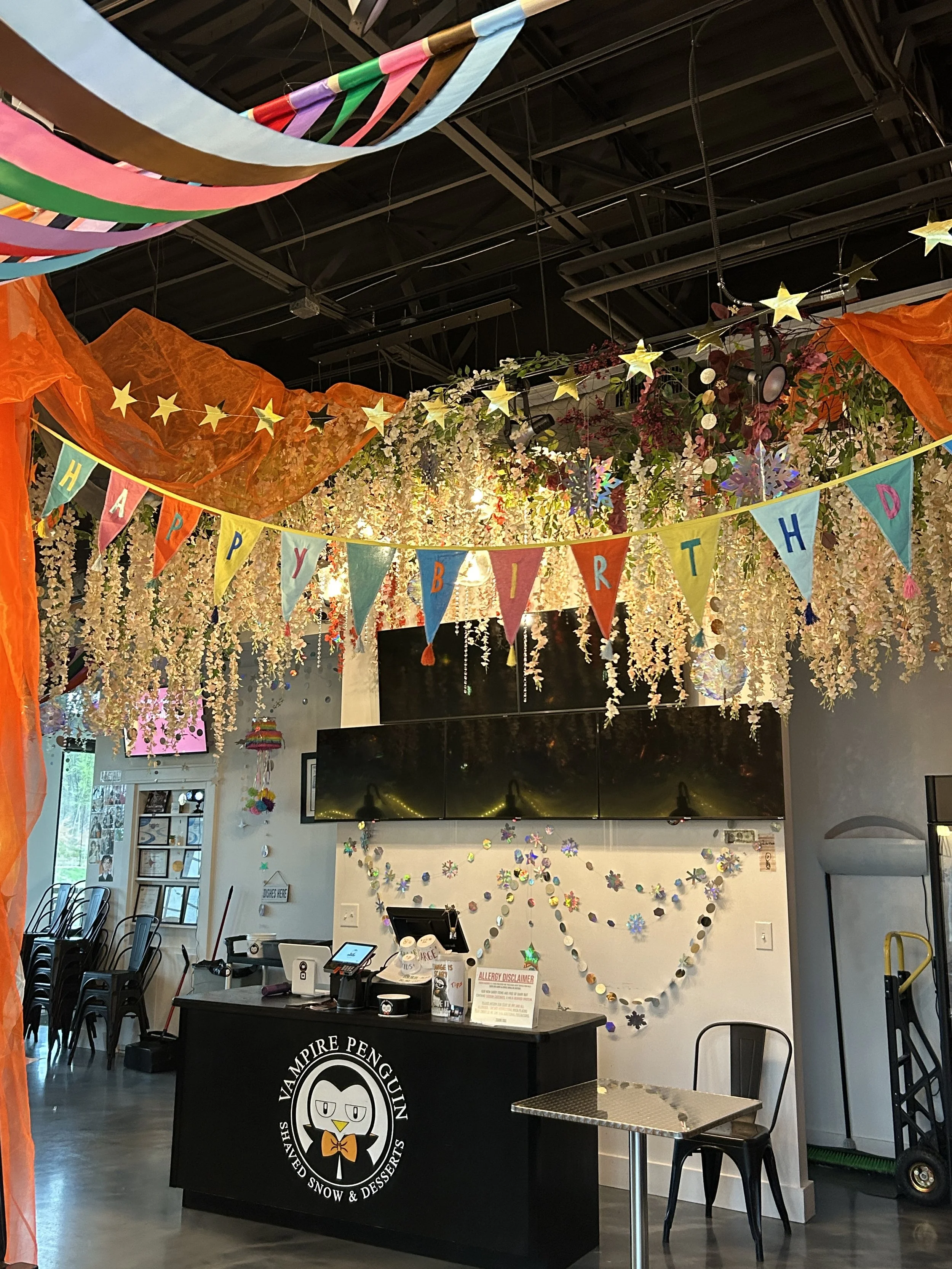 Decorated indoor space with a birthday celebration theme, featuring colorful banners spelling 'Happy Birthday,' hanging gold stars, floral arrangements, and a counter with a penguin logo that reads 'Vampire Penguin Shaved Snow & Desserts.'