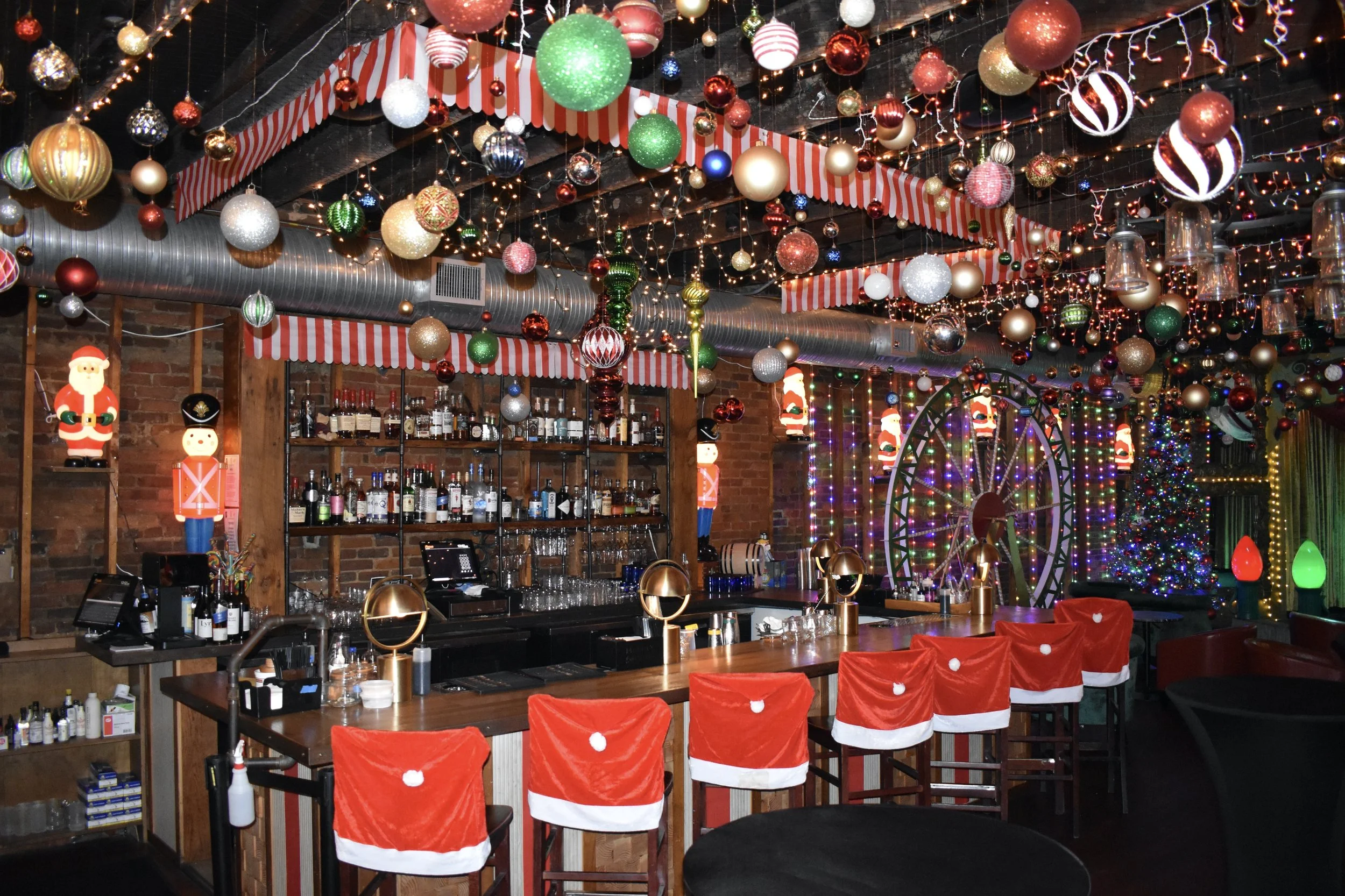 A bar decorated for Christmas with ceiling ornaments including baubles, string lights, and festive decorations. Chairs are draped with Santa hats, and a decorated Christmas tree is visible in the background.