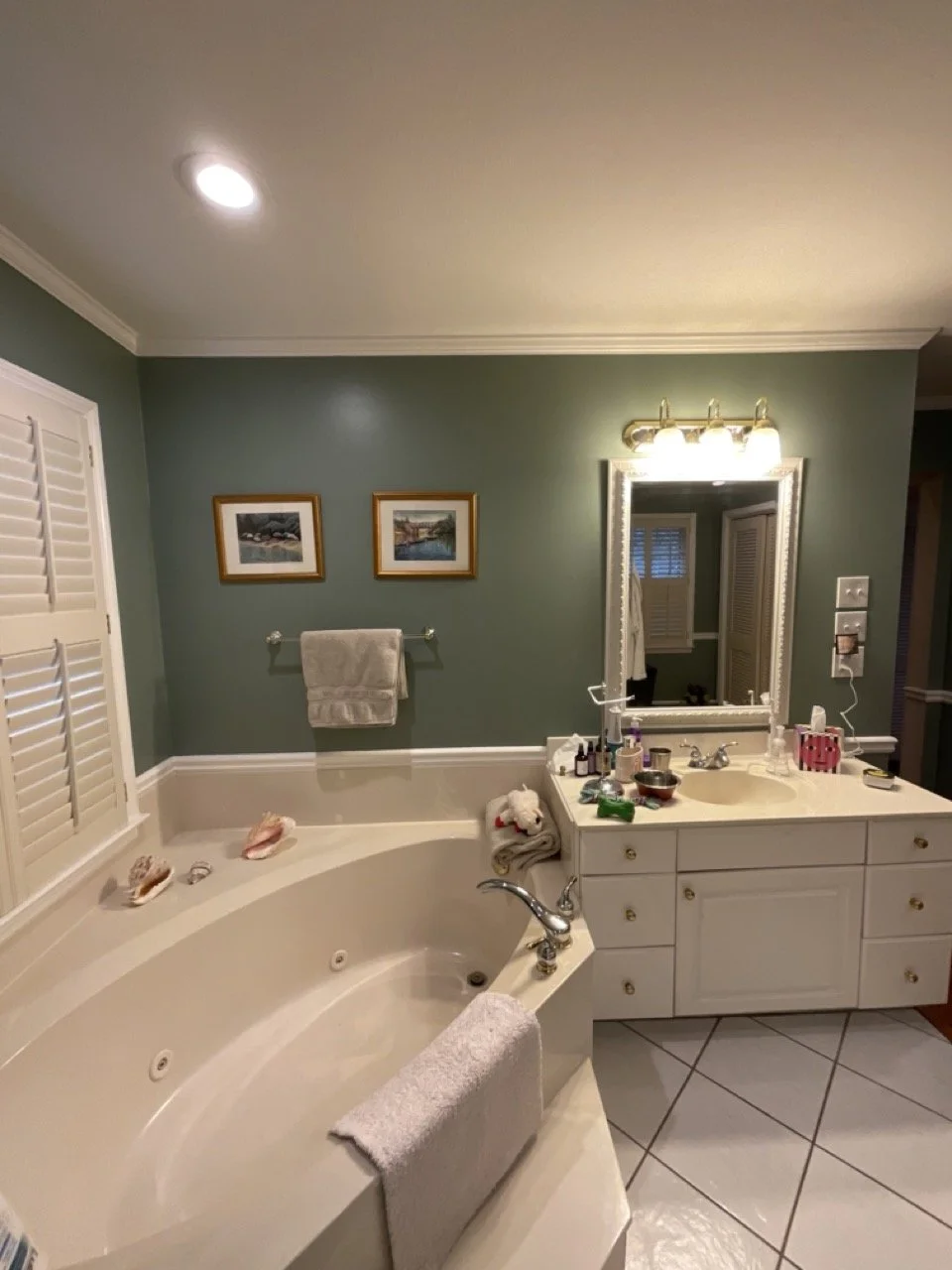 A bathroom with a corner bathtub, a white vanity with a mirror, and various toiletries on the countertop. There is a towel on the bathtub and two framed pictures on the wall above it. The room has green walls and white tiled floor.