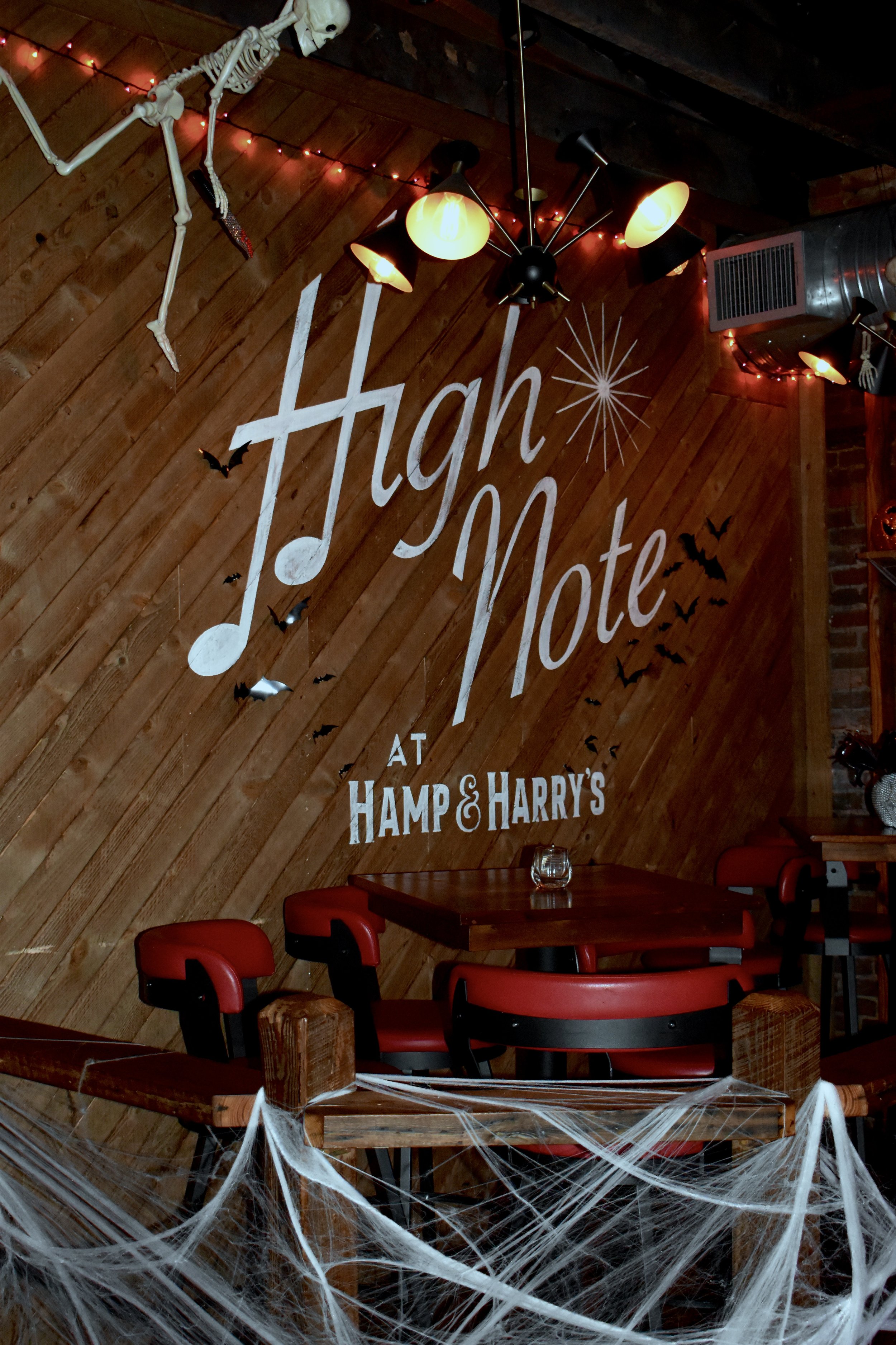 Halloween decorations inside a restaurant or bar, including a skeleton hanging from the ceiling, black bats, spider webs, and Halloween-themed signage on a wooden wall reading 'High Note at Hamp & Harry's'.