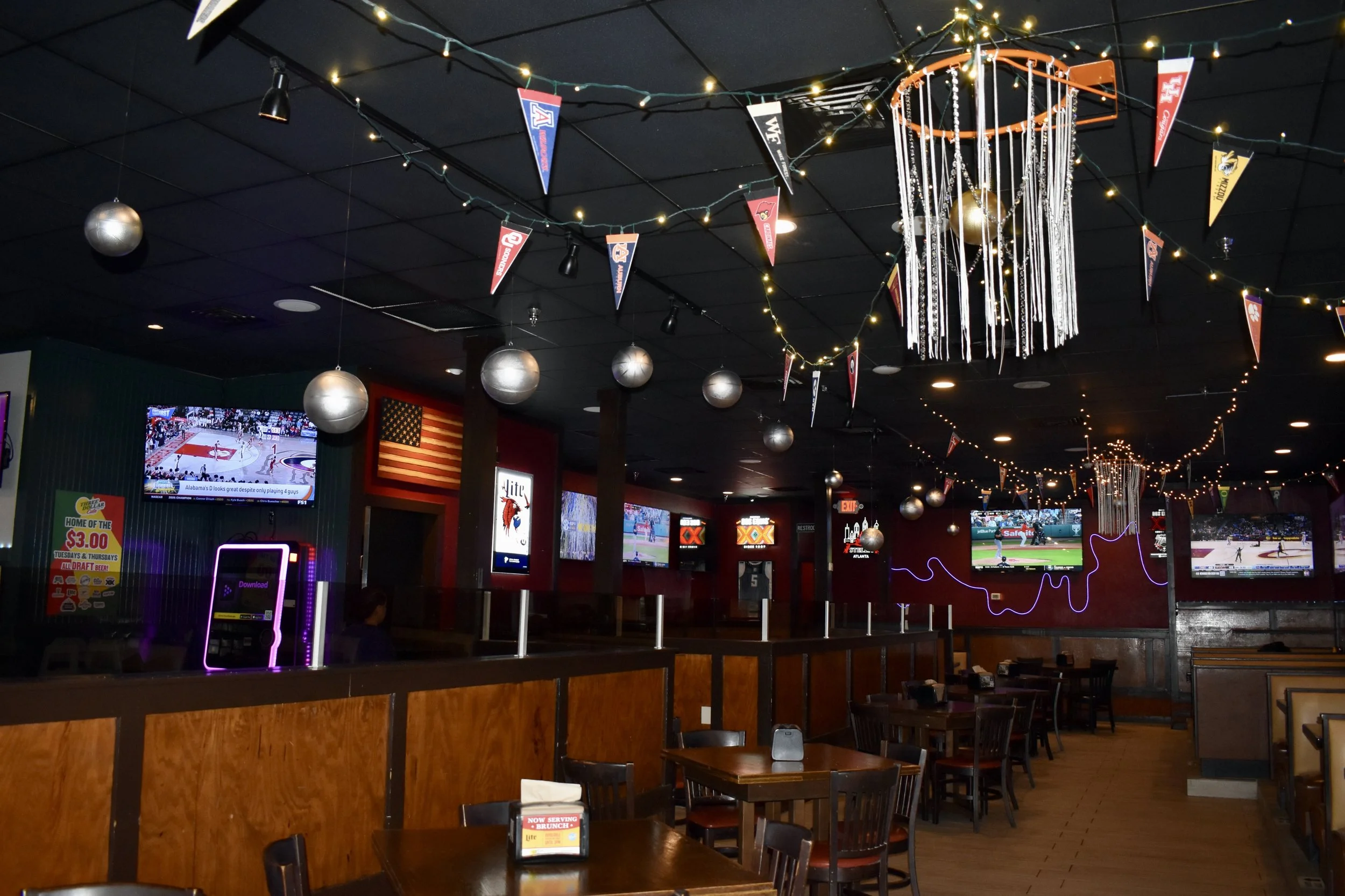 The image shows the interior of a sports bar decorated with string lights, pennant banners, and silver balls hanging from the ceiling. Multiple televisions display sports games, and there are wooden tables and chairs arranged throughout the space. 
