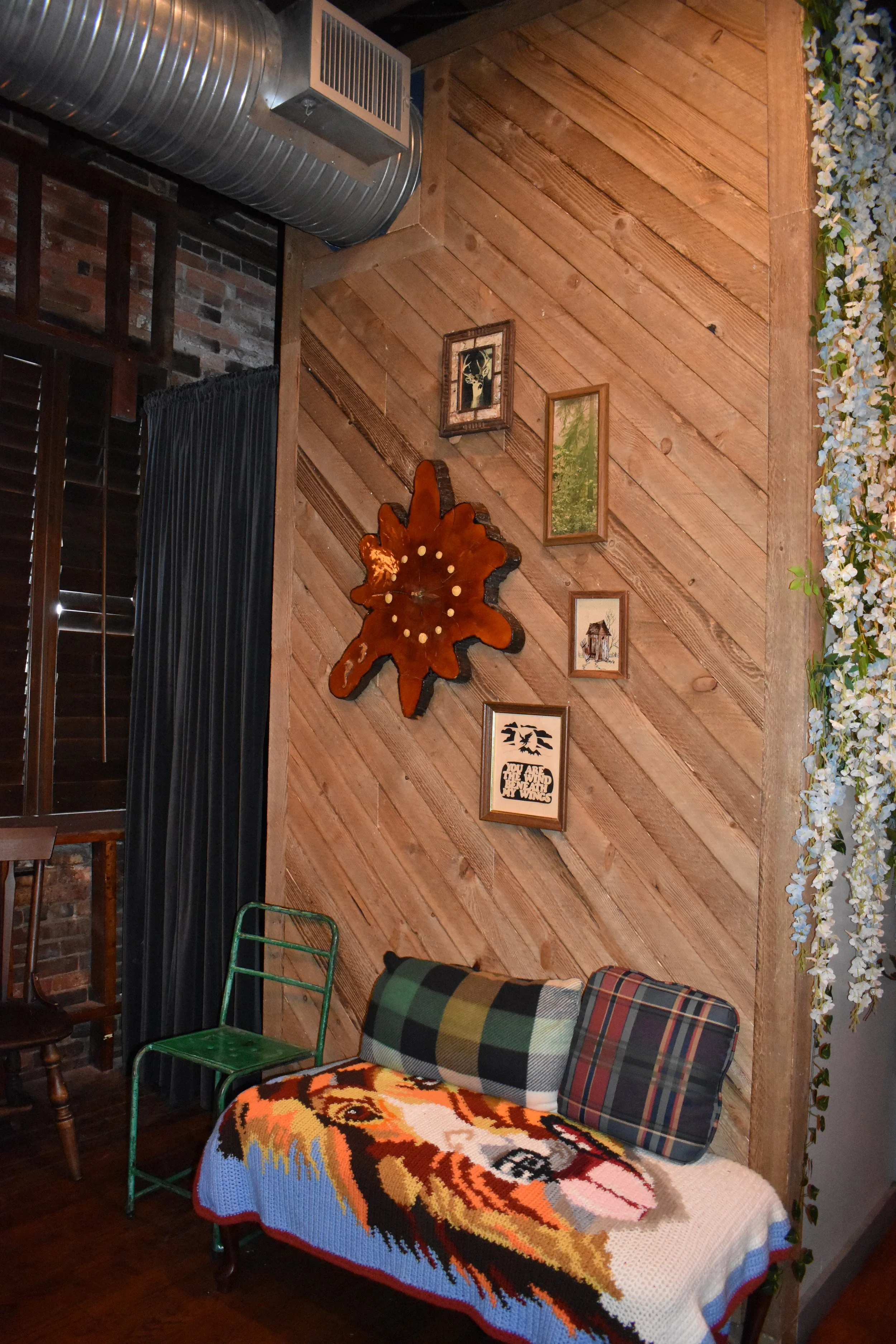 Decorative wall with framed pictures and a wooden clock, next to a window with black curtains, green metal chair, and a woven couch with a colorful afghan and pillow, in a rustic interior. Hamp & Harry's Marietta Square.