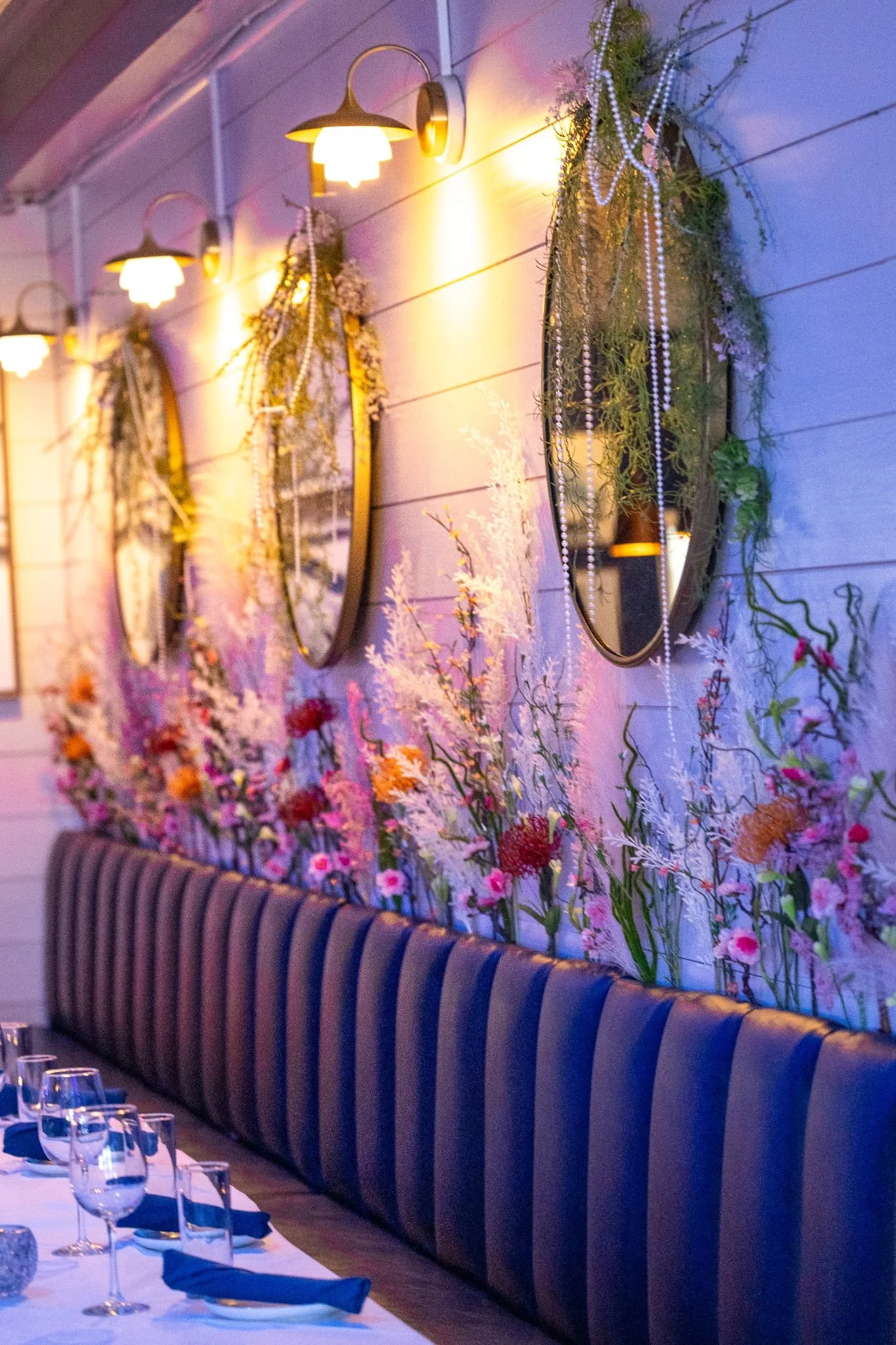 Decorative wall with multiple mirrors adorned with greenery and beads, illuminated by warm wall-mounted lights, and a long dining table set with wine glasses and blue napkins.
