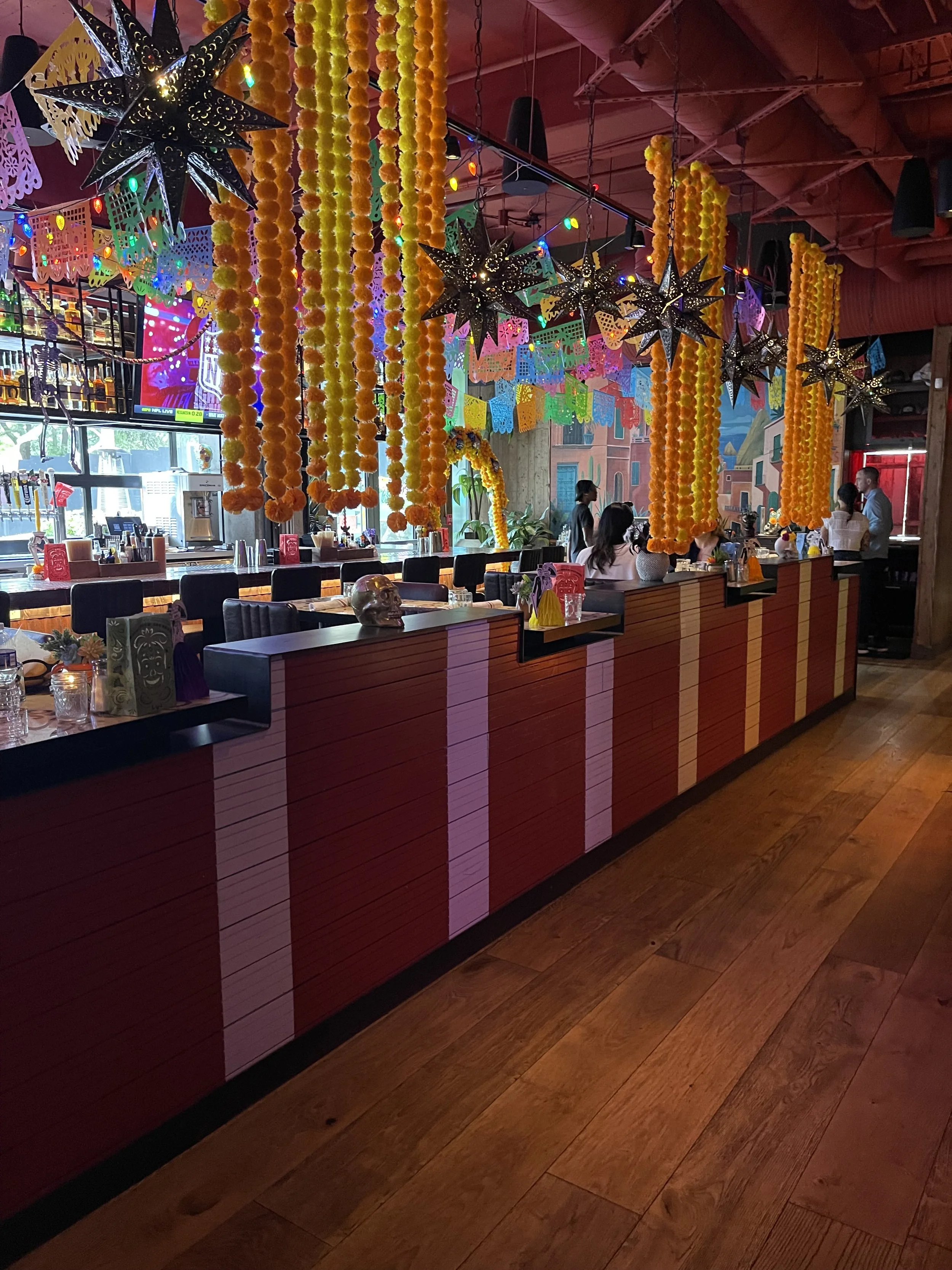 Interior of a restaurant decorated for a celebration, featuring hanging yellow and orange paper garlands, black star-shaped lanterns, colorful papel picado banners, and a wooden bar or counter with chairs and tables. Emilio's Tacos & Tequila Midtown 