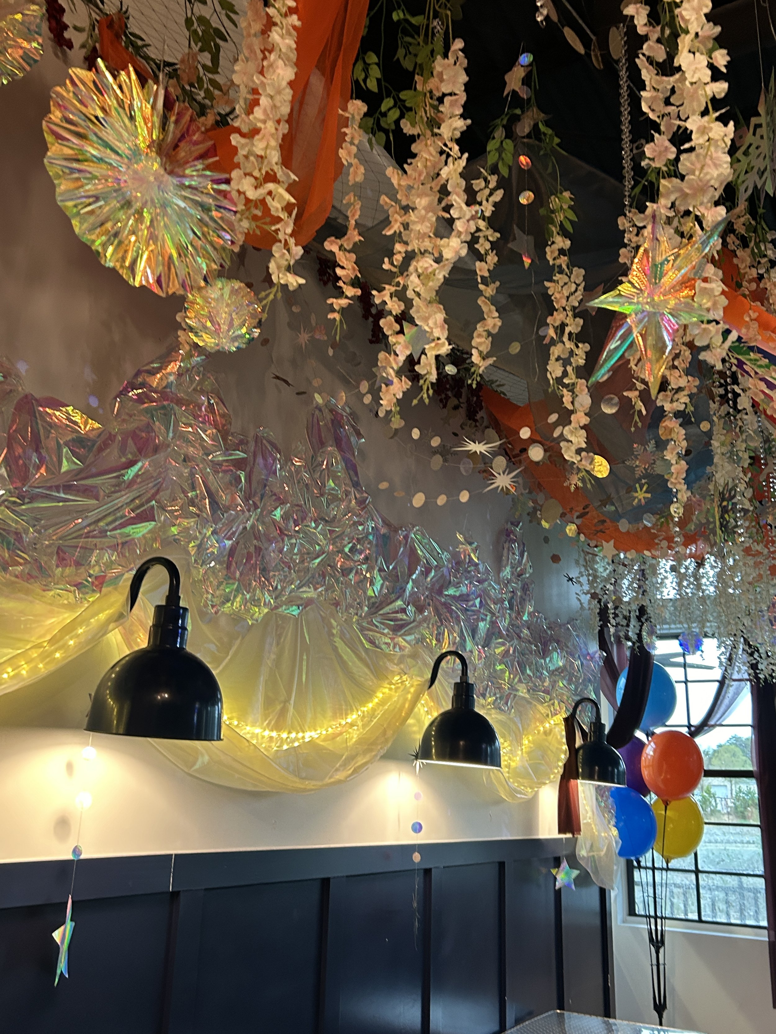 Decorative indoor ceiling setup with hanging pink and white flowers, holographic foil streamers, star and circular hanging ornaments, black wall sconces with warm yellow lighting, yellow tulle fabric and string lights, and colorful balloons in the co