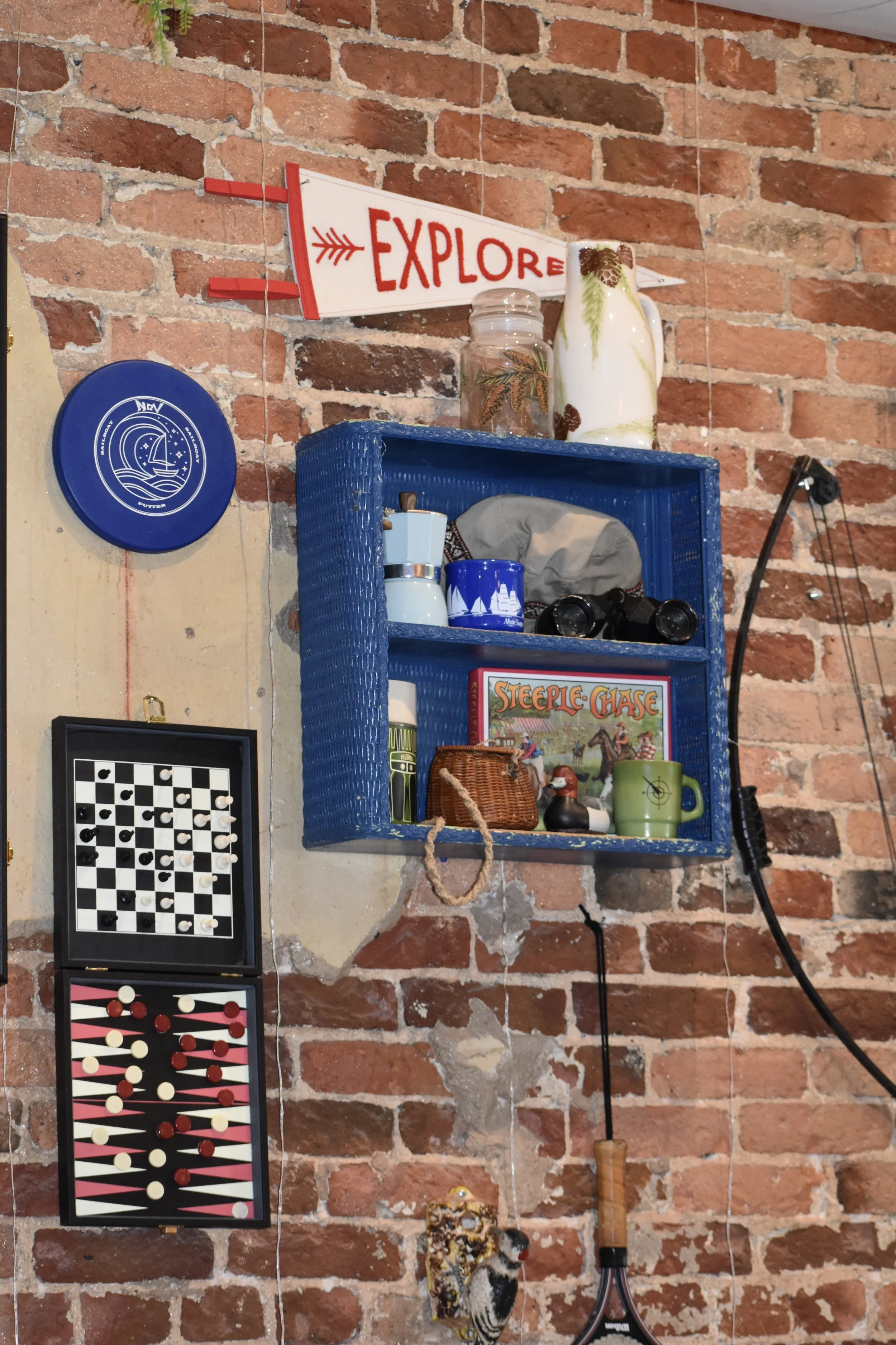 Decorative items on a rustic brick wall including a blue basket shelf with mugs, a board game, and small figurines, with a sign that says 'Explore' and a fishing rod. Hamp & Harry's Marietta Square.