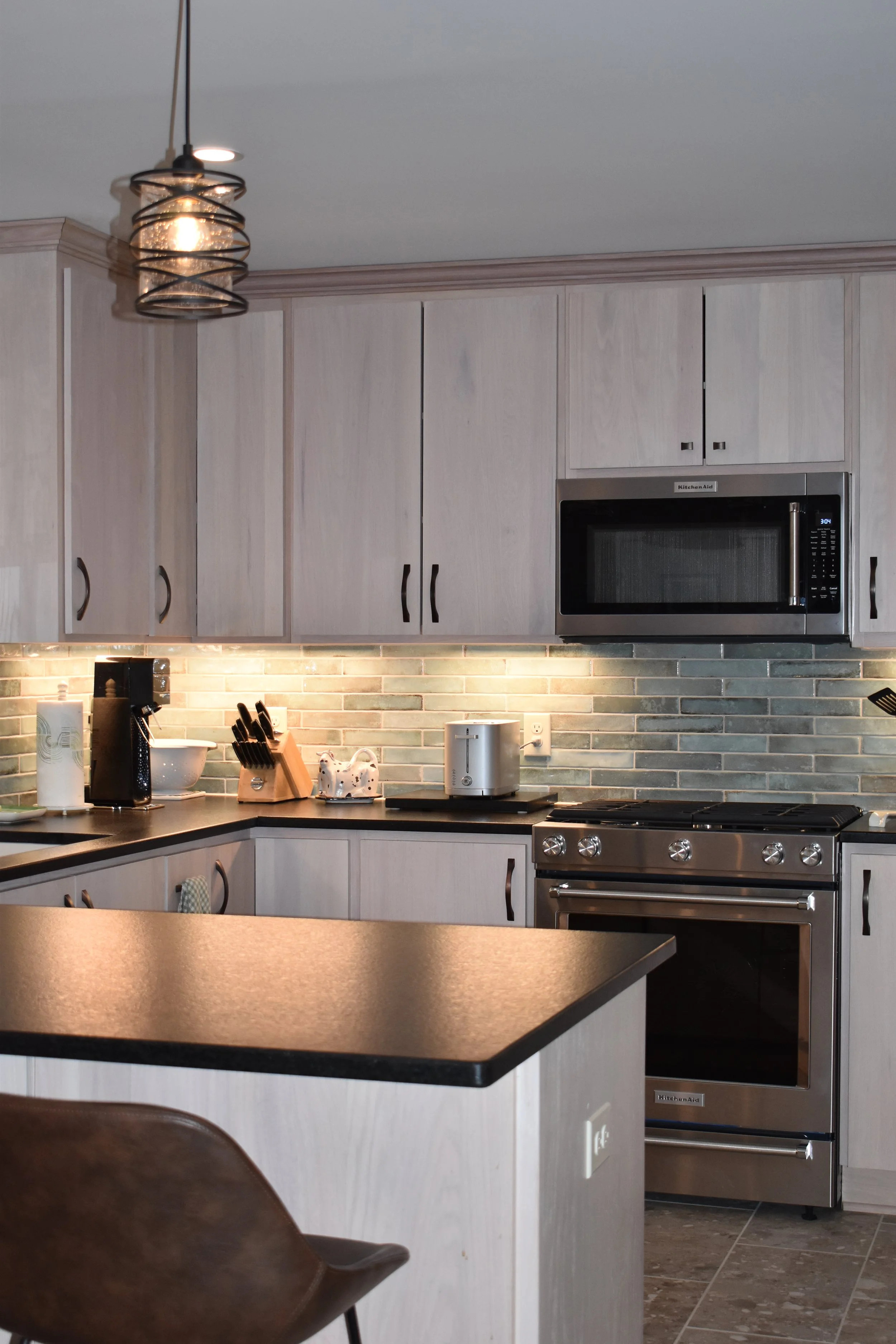A modern kitchen with light-colored wooden cabinets, a black countertop, stainless steel appliances including a microwave and oven, and a brick-style backsplash. There is a paper towel roll, a coffee maker, a knife block, and a toaster on the counter