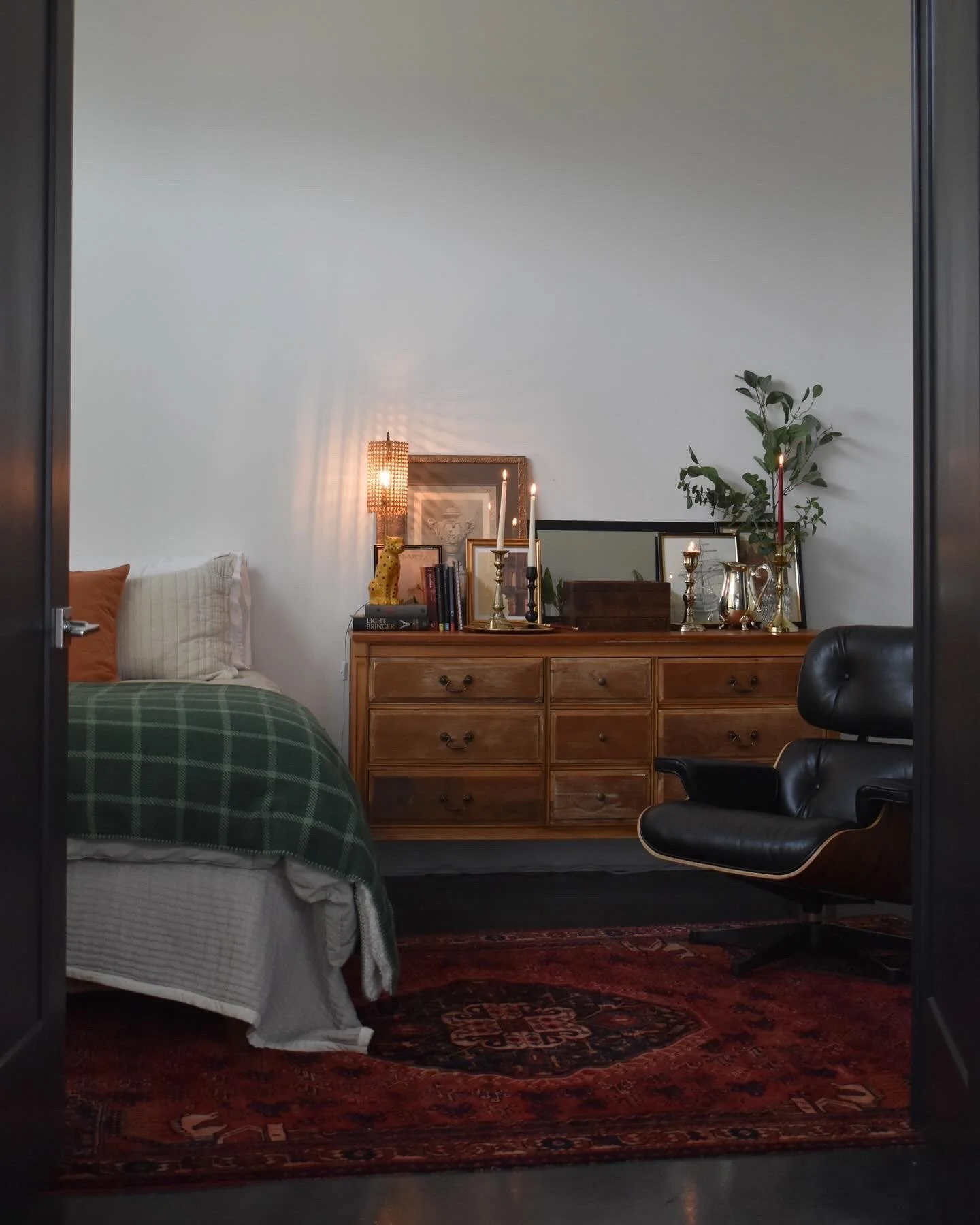 A cozy bedroom with a bed on the left covered in a green plaid blanket and pillows. A wooden dresser on the right holds framed art, candles, books, and decorative items, with a tall plant behind it. An Eames lounge chair is to the right of the dresse