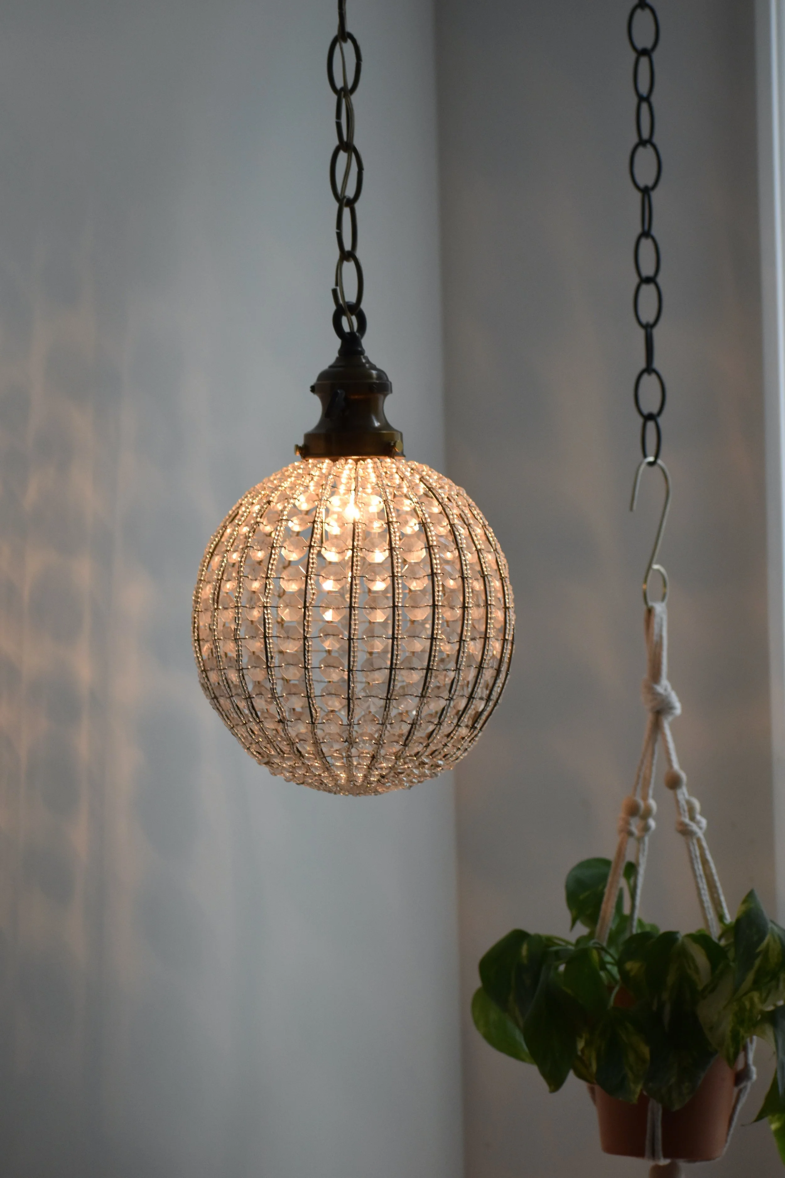 Hanging decorative light fixture with a round, crystal-like exterior, casting light and shadows on the wall behind it, next to a potted plant in a hanging planter.