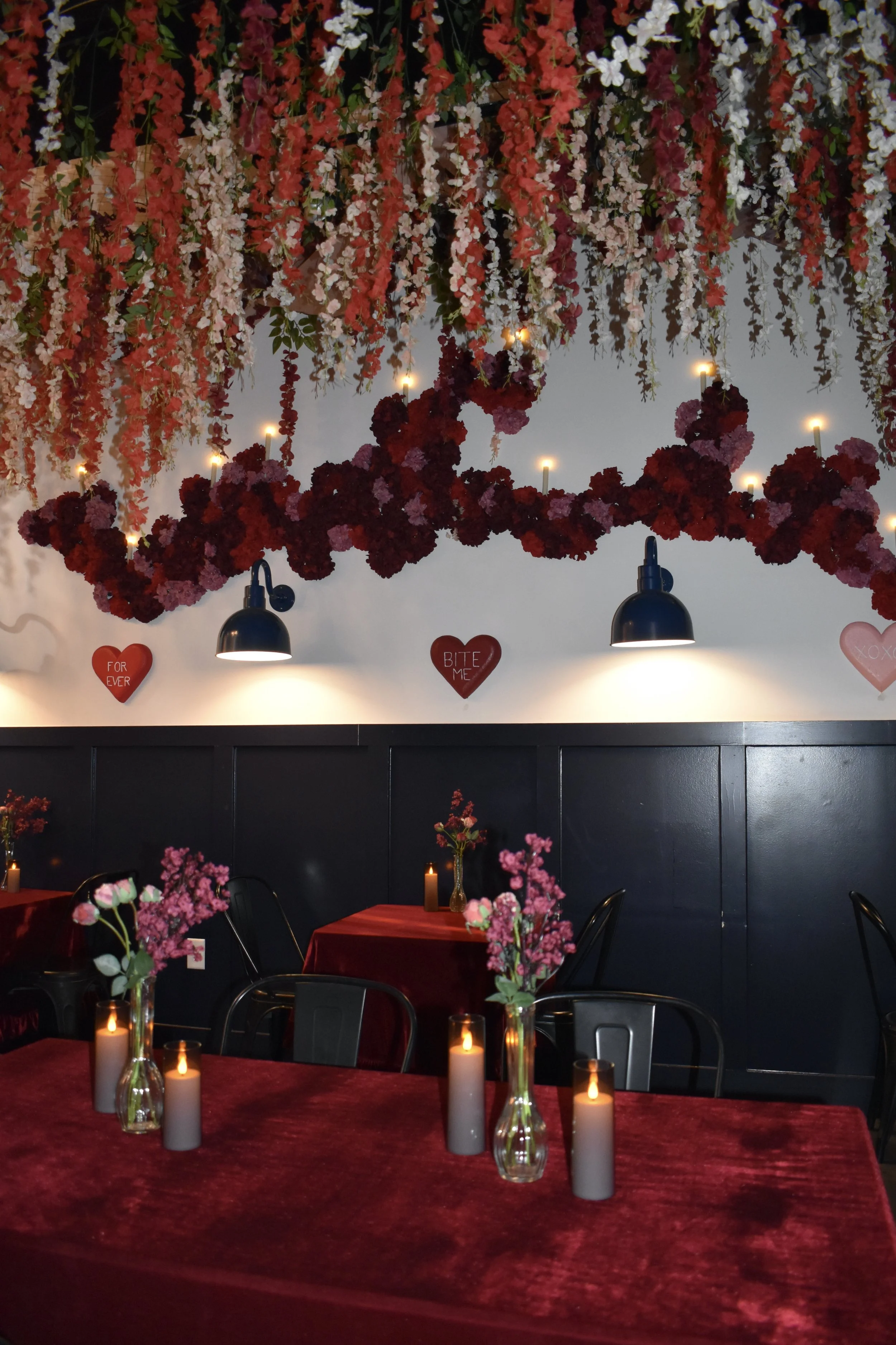 A romantic decorated dining area with hanging flower arrangements in shades of pink, red, and white, black wall panels, black hanging lamps, pink and red flowers in vases, and candles on red velvet tablecloths. Wall signs with love-themed messages.