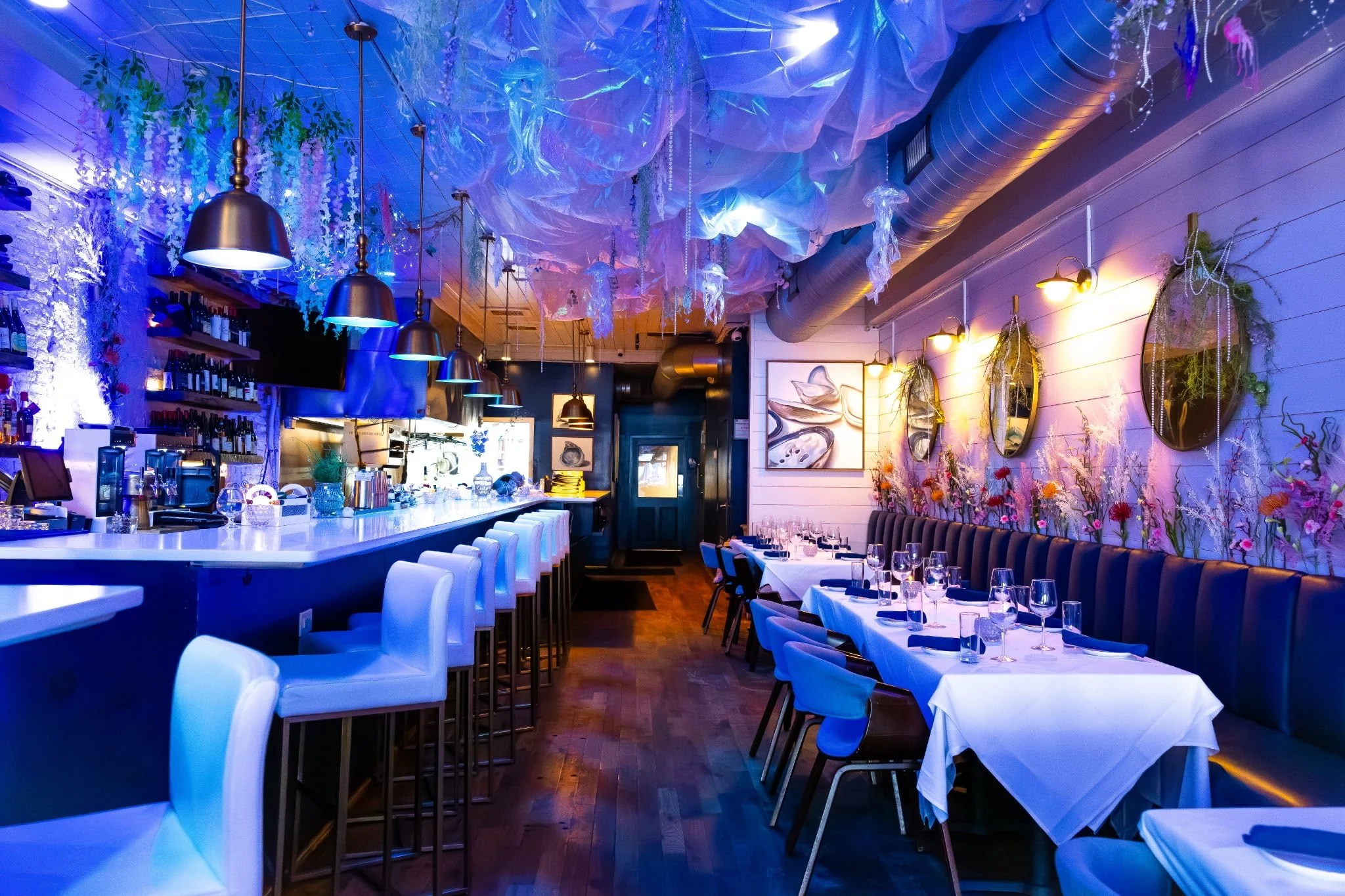 Upscale restaurant interior with white bar counter, high white chairs, and a dining area with tables set with wine glasses and tableware. Decor includes floral arrangements, hanging mirrors, and artwork, with purple and blue ambient lighting and deco