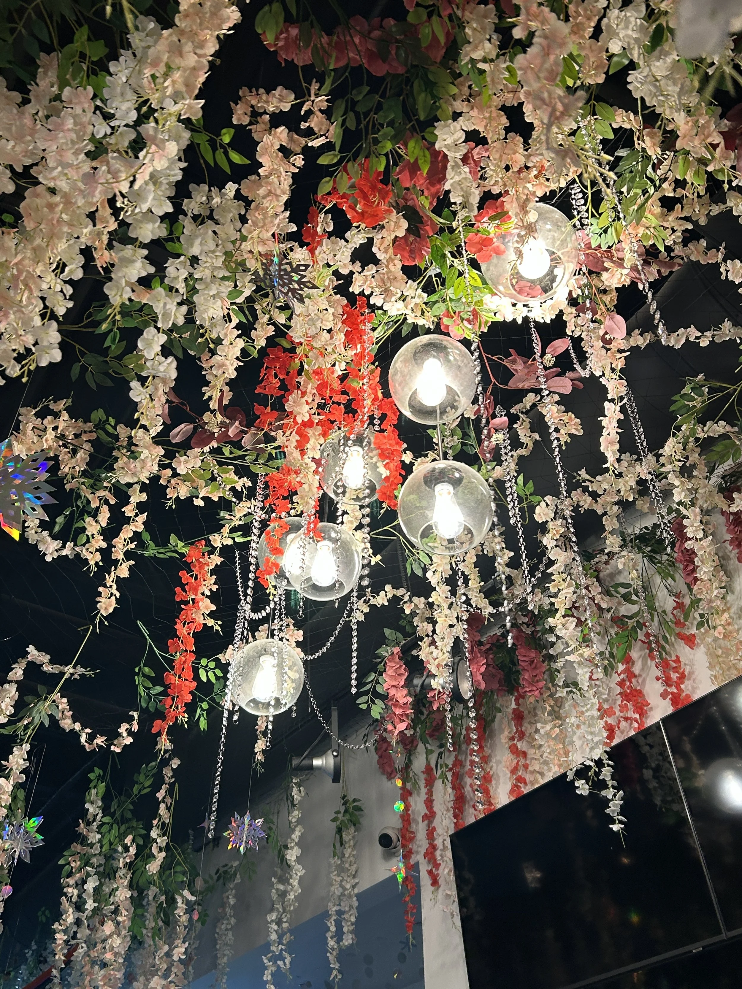 Ceiling decorated with pink, white, and red artificial flowers, hanging glass bulb light fixtures, and sparkling chains.